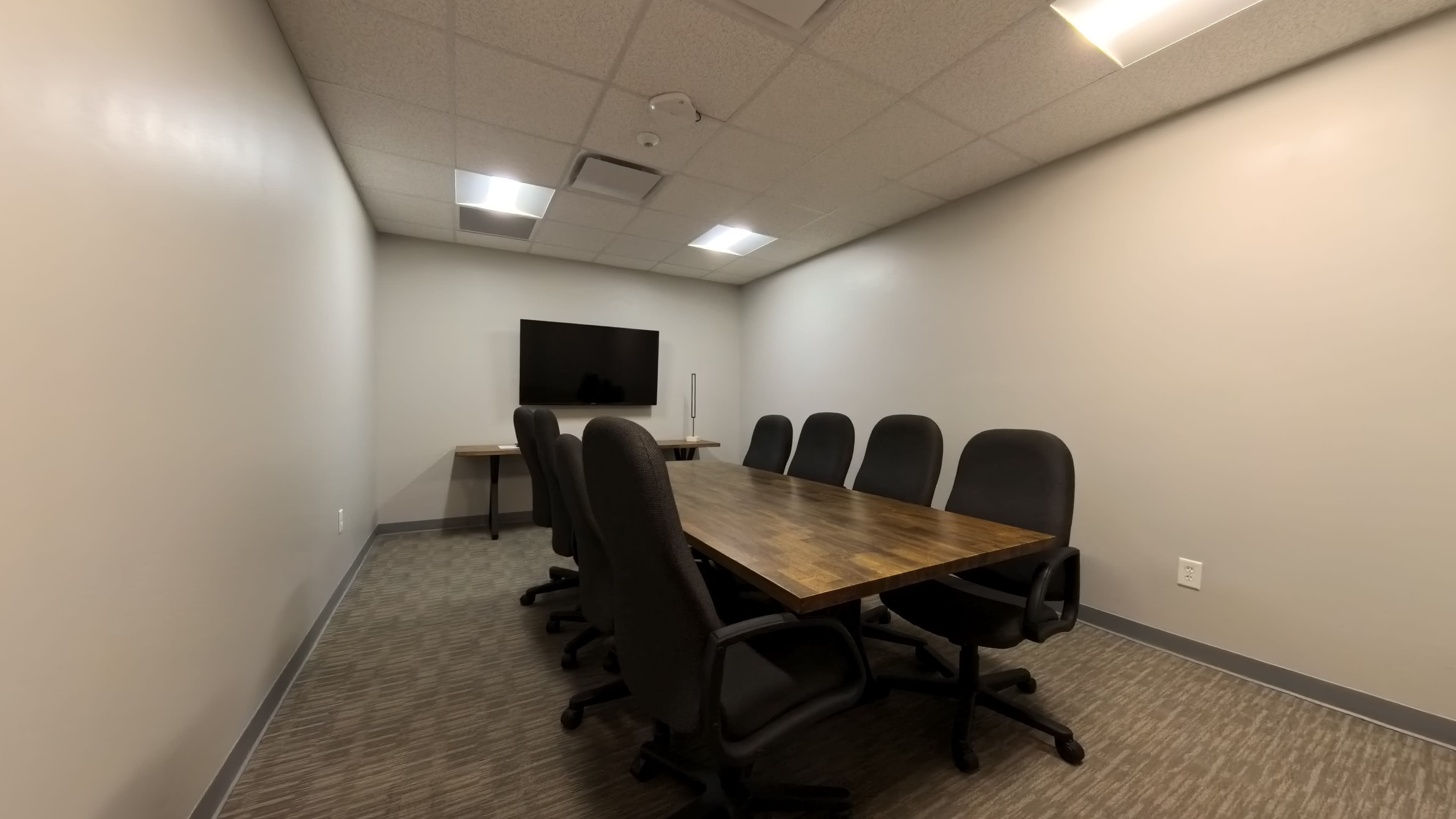 A conference room features a large wooden table surrounded by eight black chairs, with a television mounted on the wall.