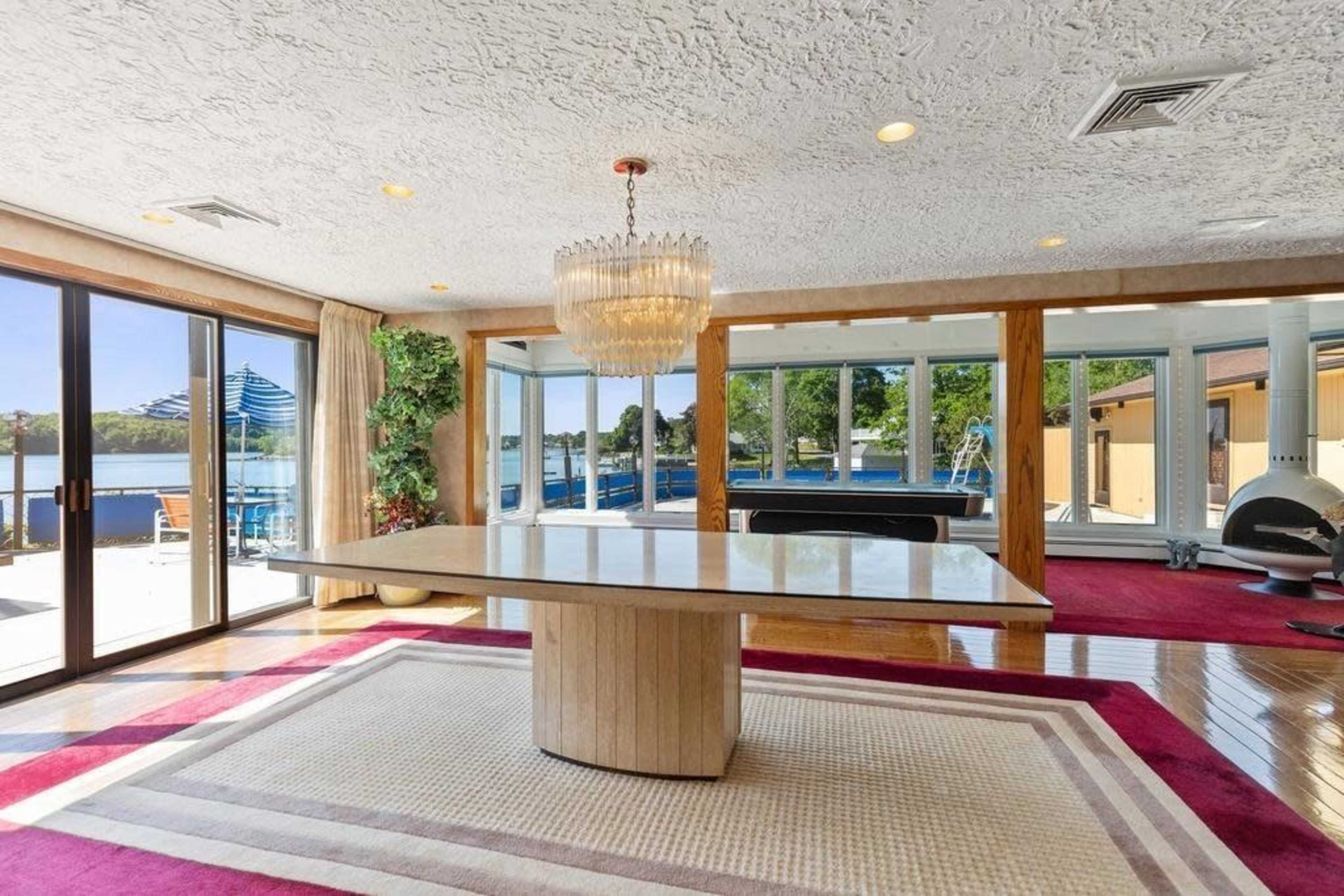 The image shows a spacious interior with a large round table in the center, overlooking a lake through large glass doors.
