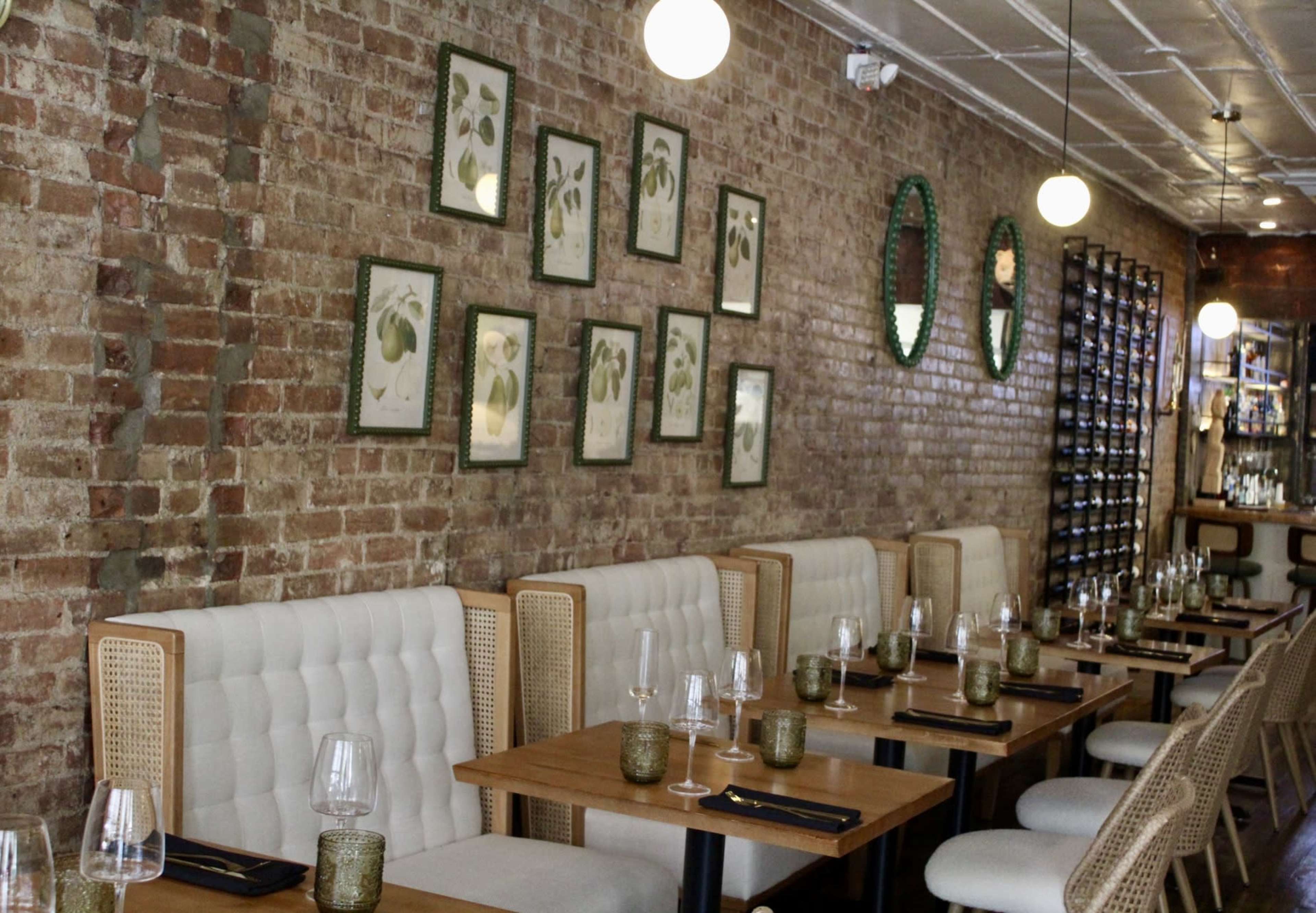 The image depicts a restaurant interior with brick walls, wooden tables set with glassware and candles, and framed botanical prints on the wall.