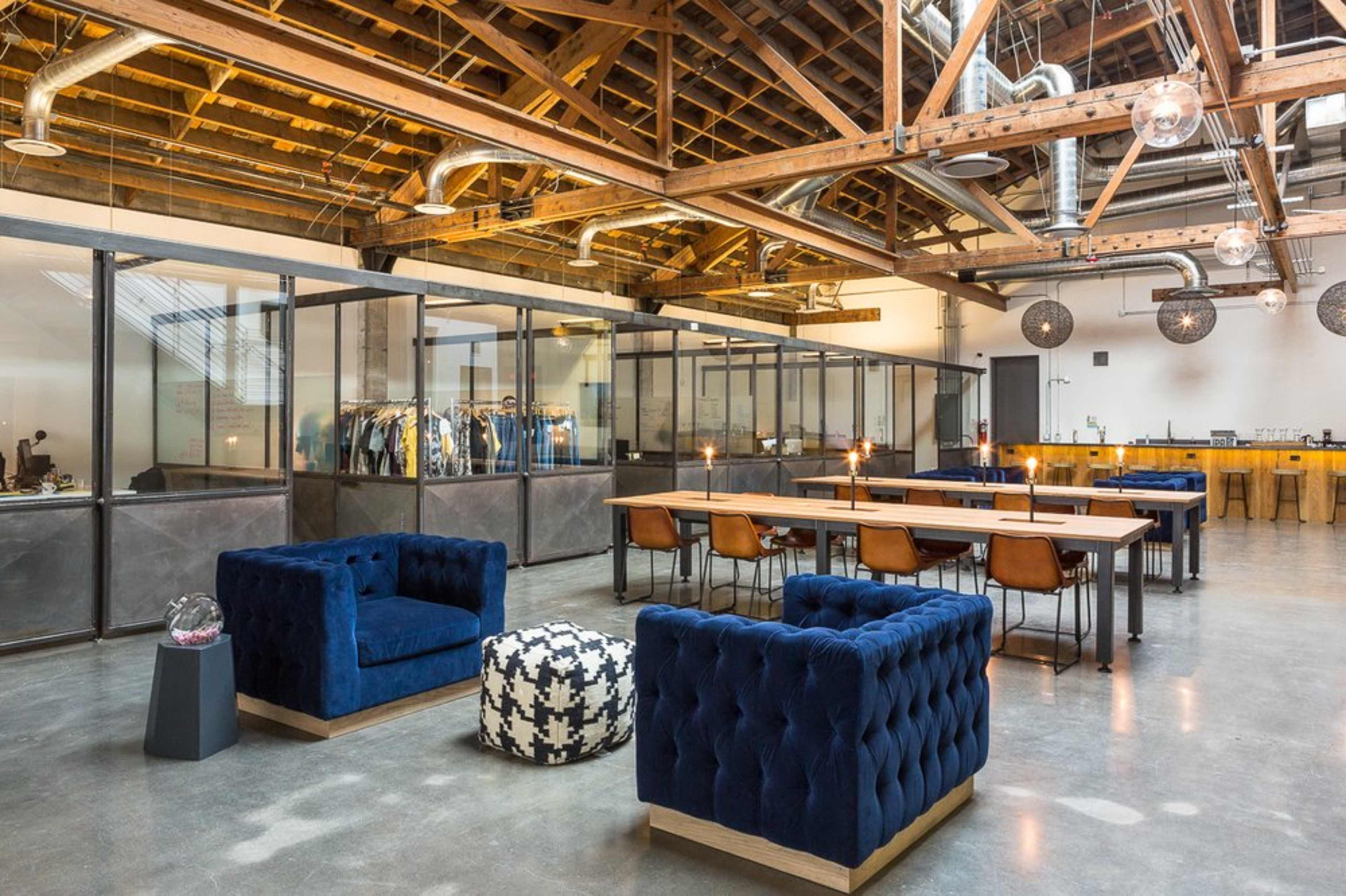 A spacious modern office with a mix of lounge seating and a large communal table, featuring exposed wooden beams and glass partitioning.