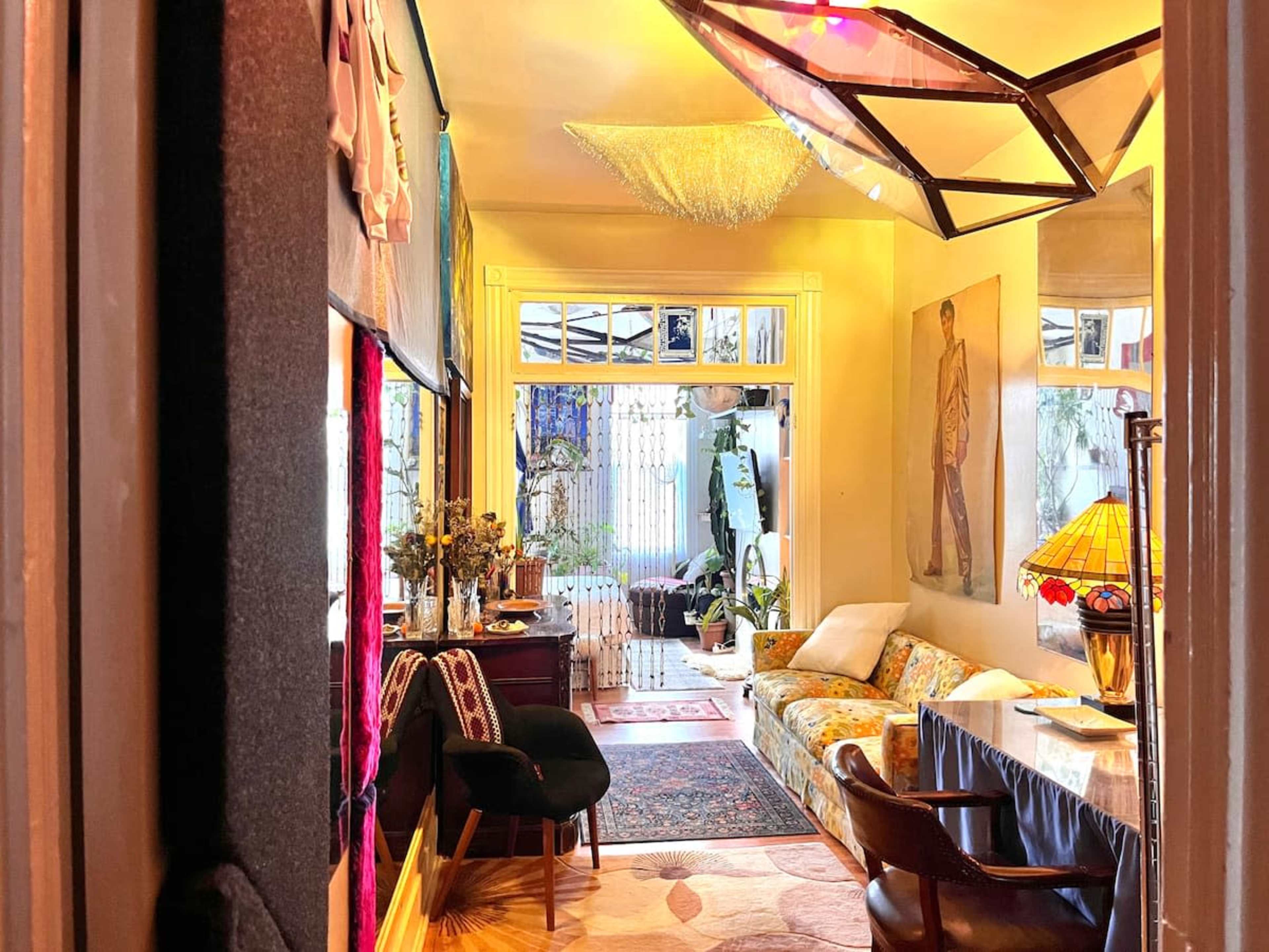 The image shows a brightly lit hallway leading to a living area with a patterned couch, a lamp, and a view of an outdoor space through large windows.