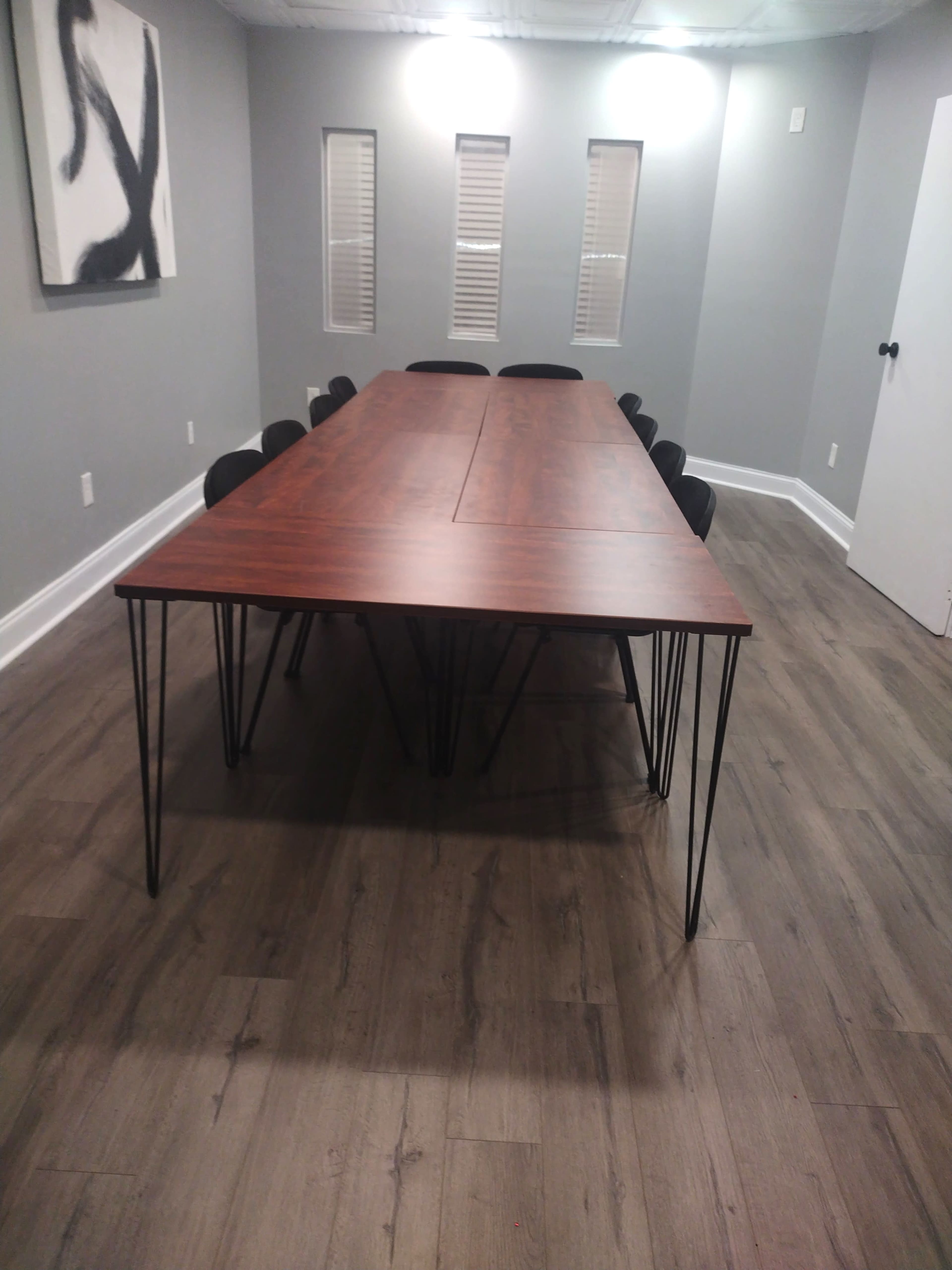 A long wooden table with metal legs is surrounded by black chairs in a well-lit, gray-walled meeting room.