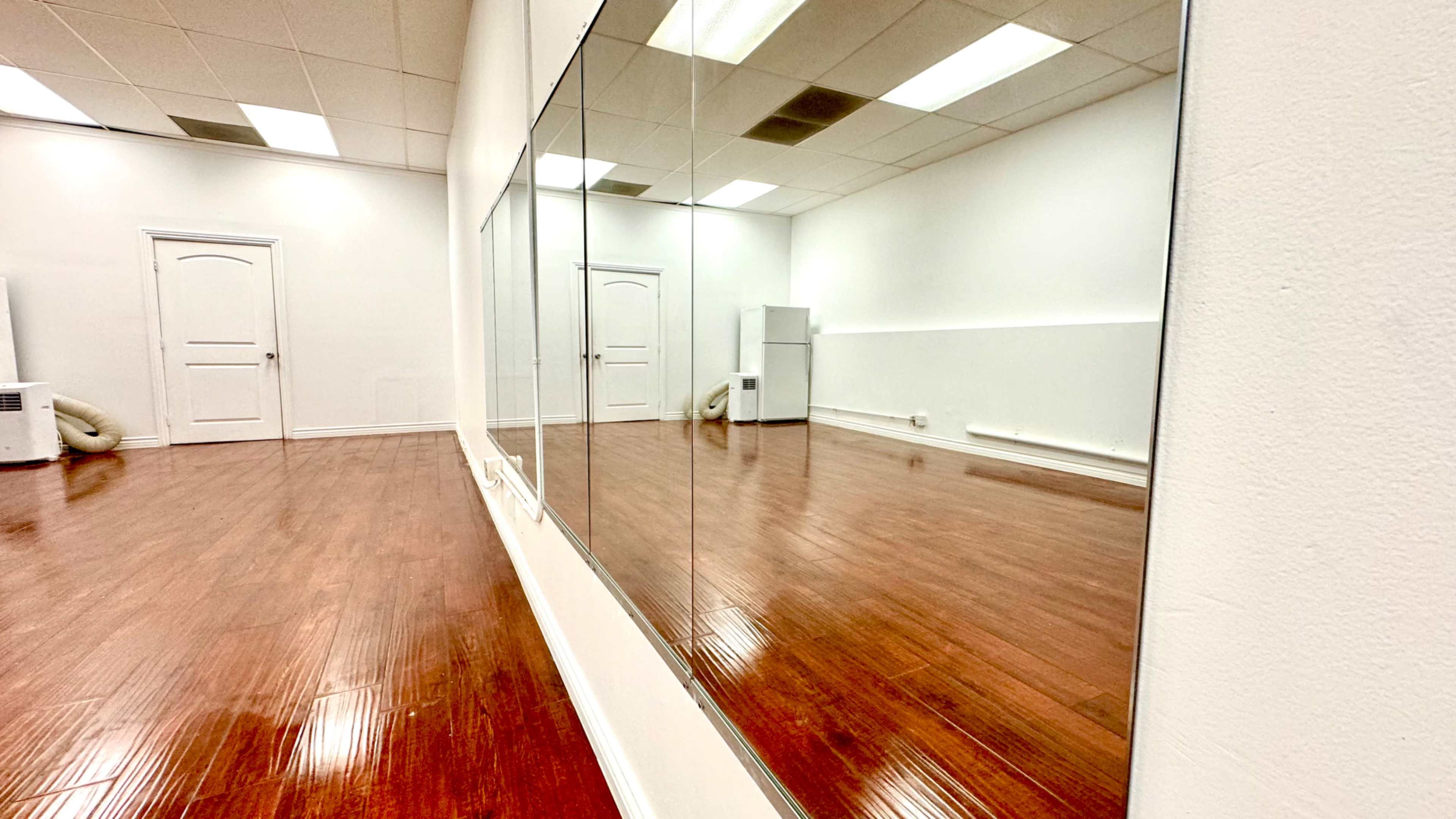 A spacious room with polished wooden floors and a wall of mirrors, along with a door and a white refrigerator in the corner.