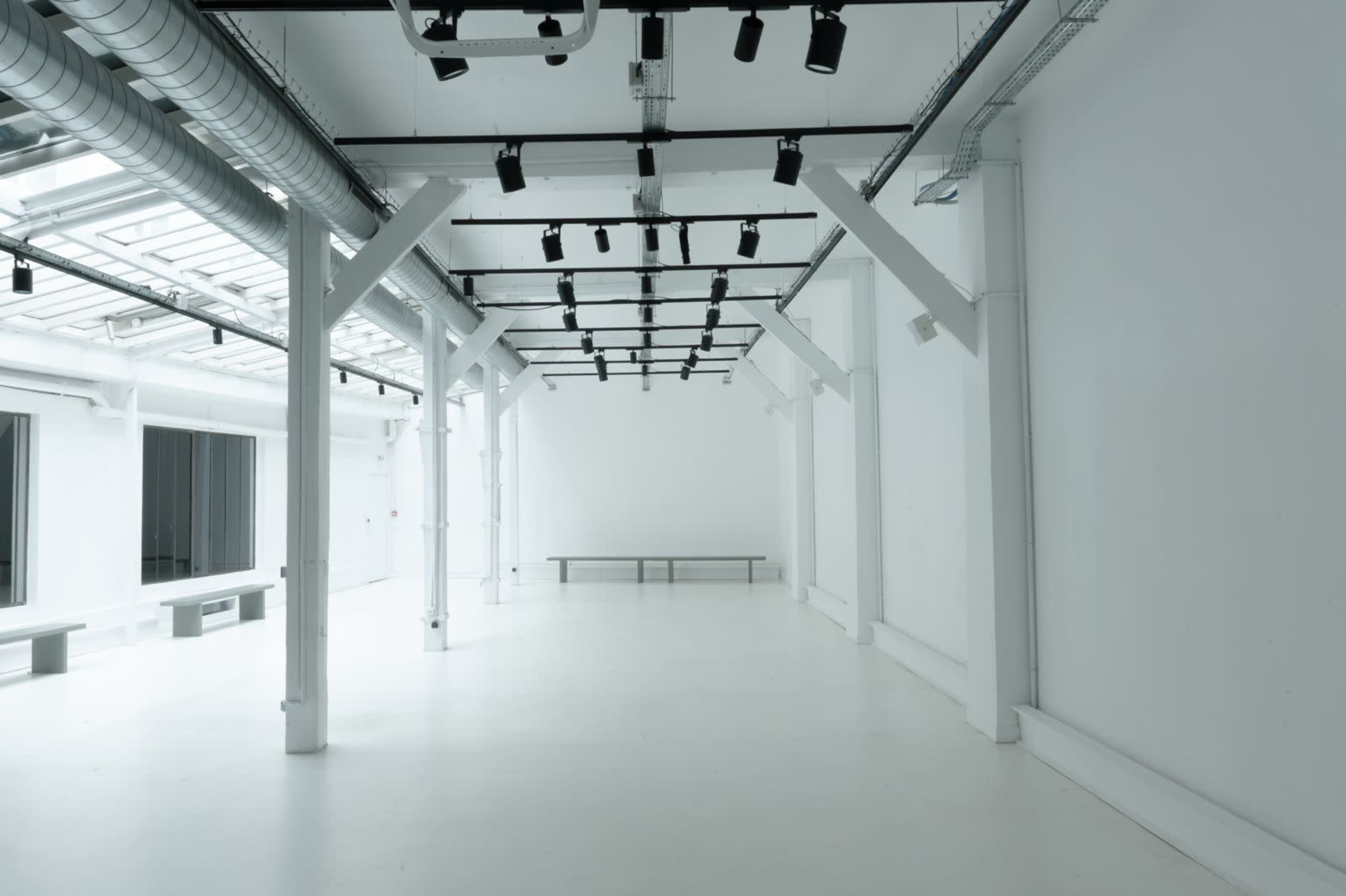 The image shows a spacious, empty white room with industrial lighting and a long bench along one wall.