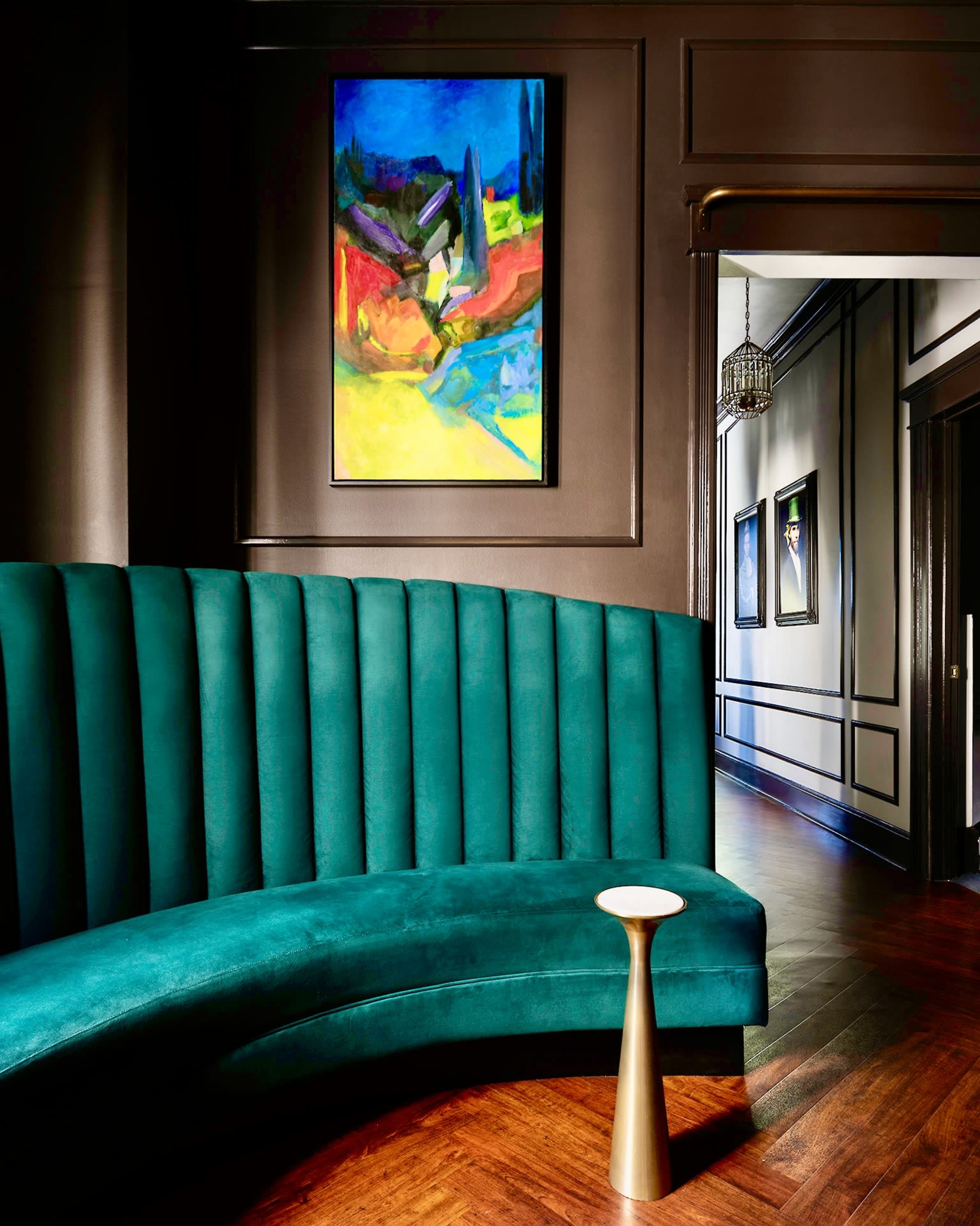 A curved teal seating area in a dimly lit room, with a colorful abstract painting on the wall and a narrow table beside it.