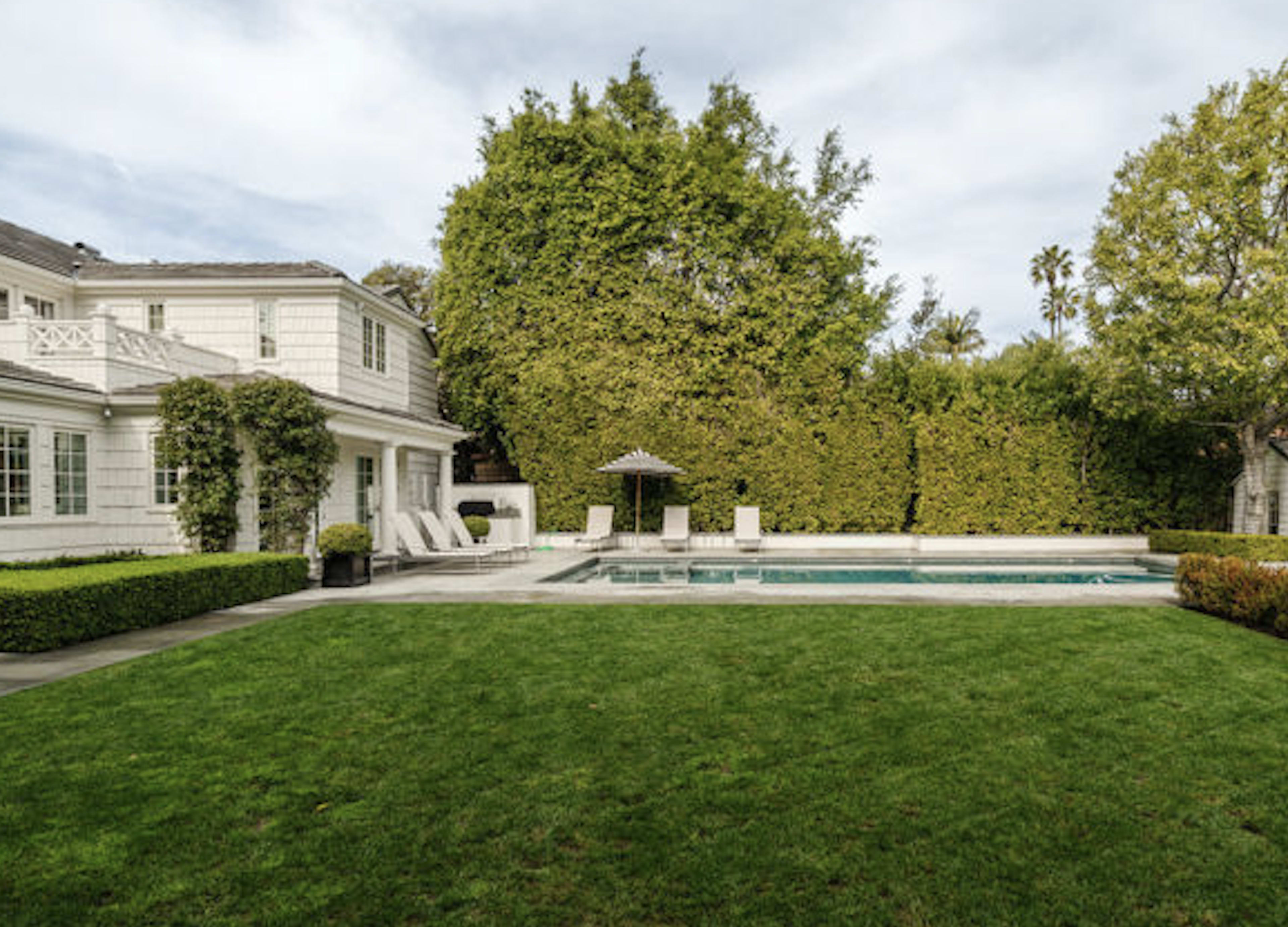 A spacious backyard features a swimming pool surrounded by lush greenery and a two-story white house.