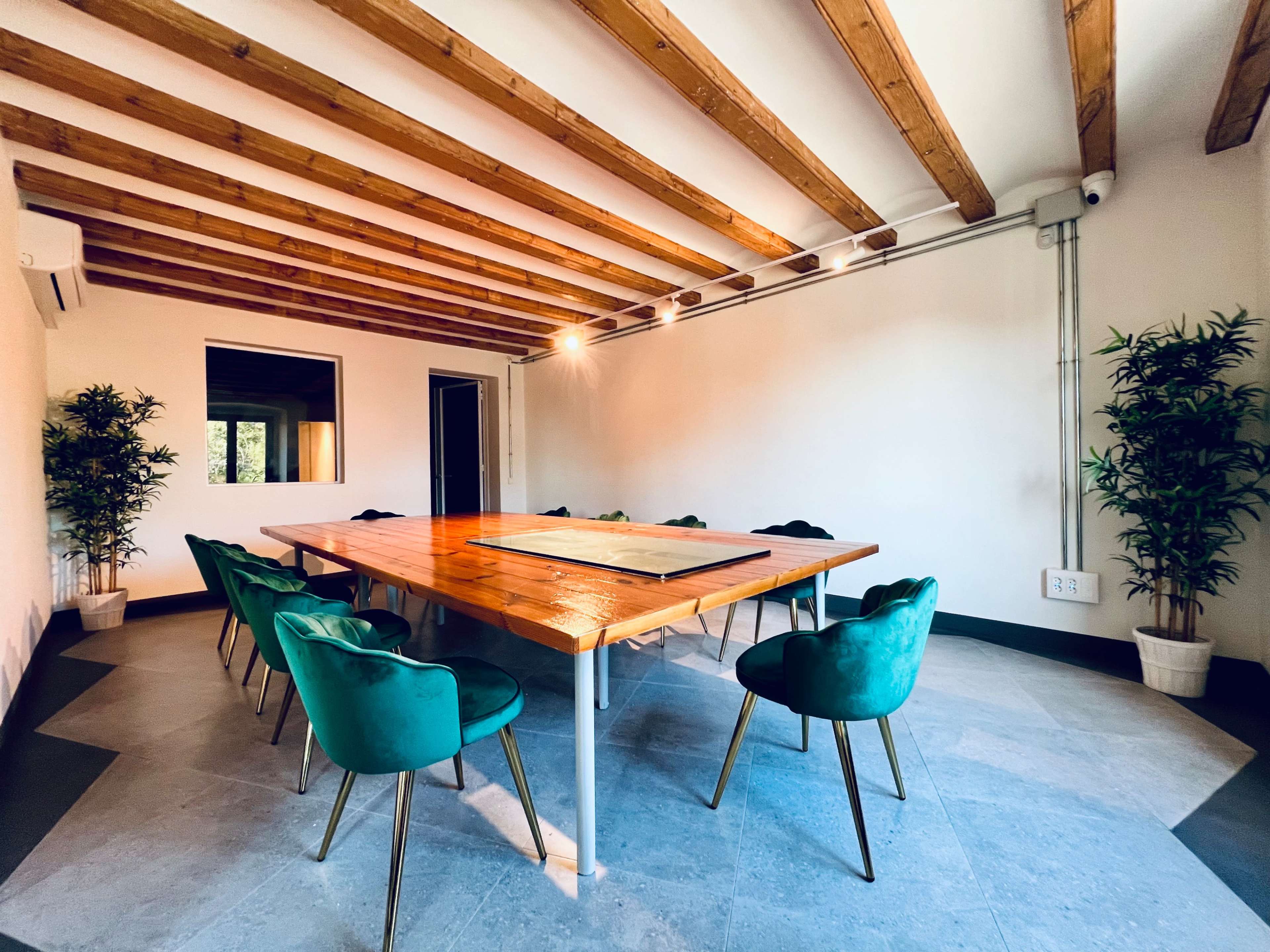 A modern conference room features a large wooden table surrounded by green upholstered chairs and small potted plants in the corners.