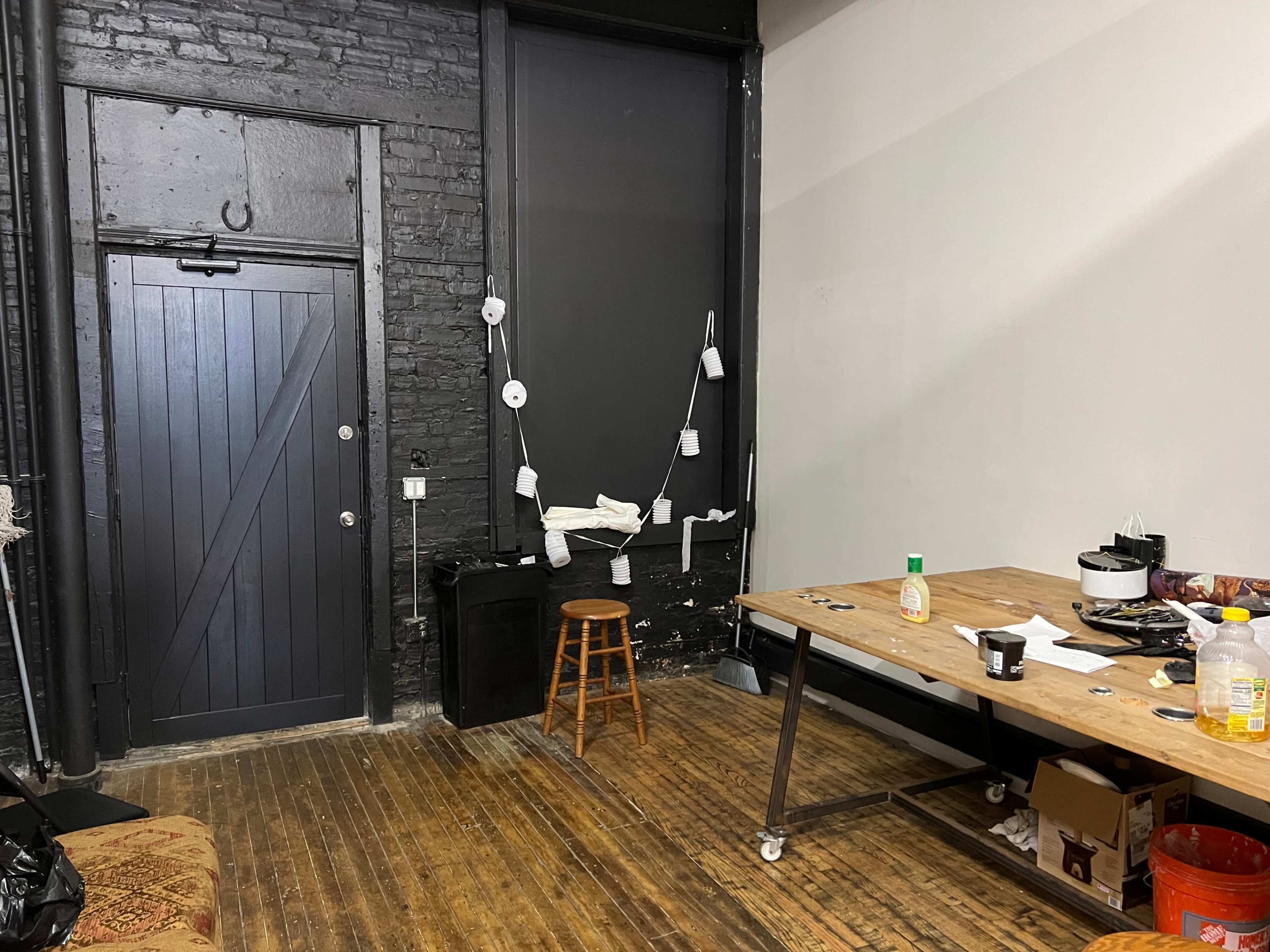 A dimly lit room with a black door, a stool, a wooden table cluttered with items, and a wall adorned with cups and paper.