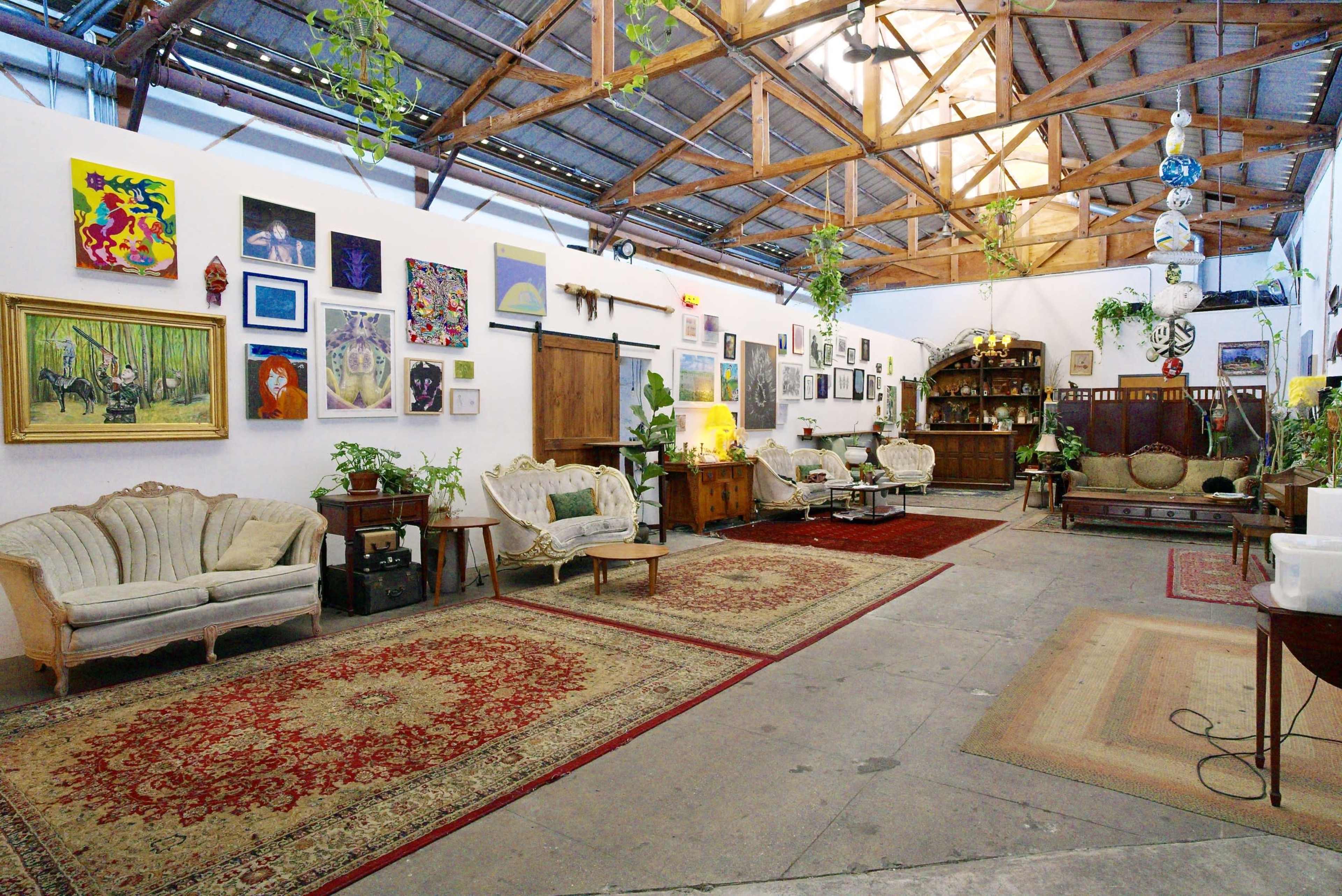 The image shows a spacious, well-decorated indoor area featuring vintage furniture, multiple artworks on the walls, and plants hanging from the ceiling.