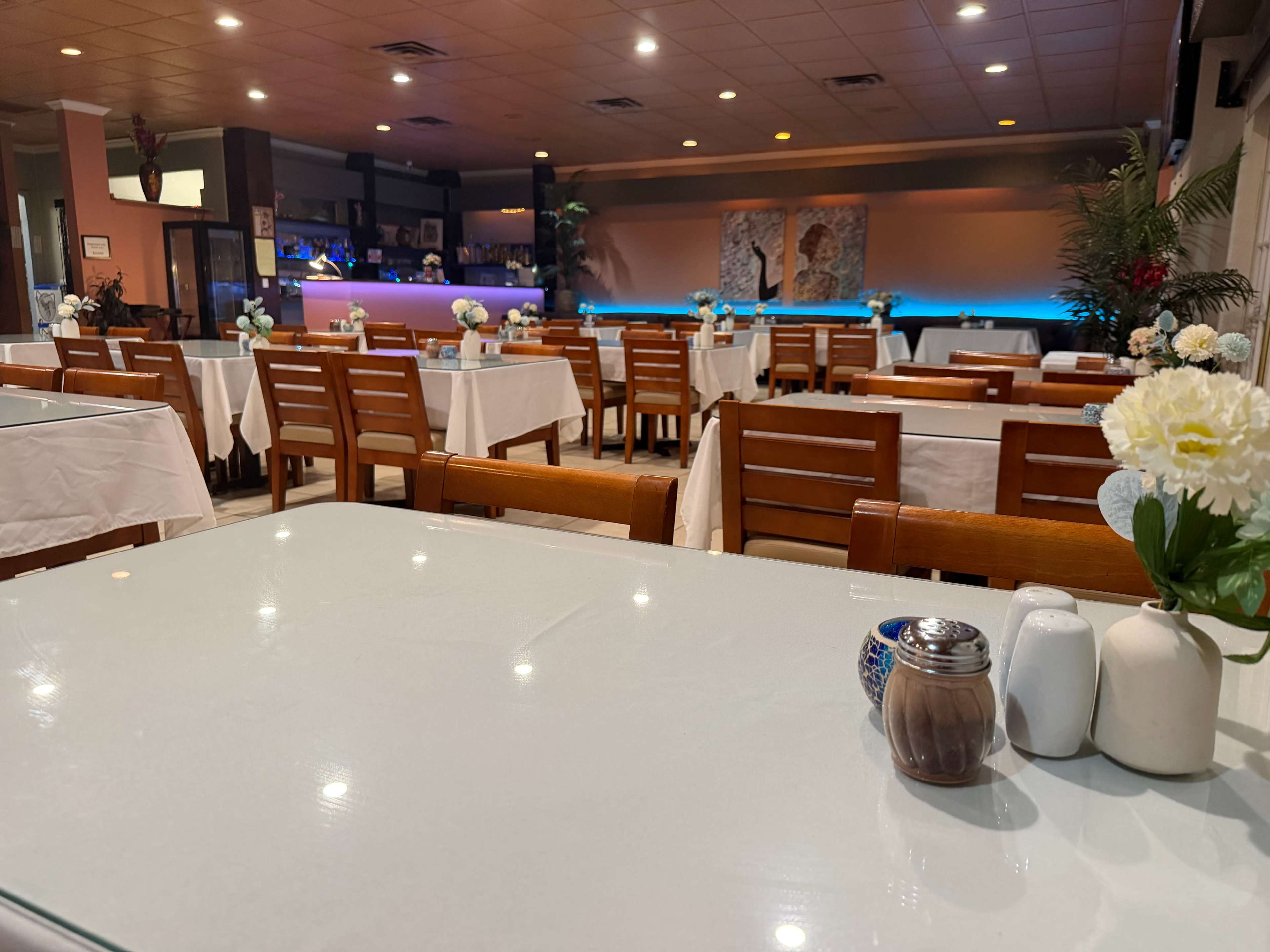 The image shows a neatly arranged restaurant interior with wooden tables and white tablecloths, featuring decorative floral centerpieces and a softly lit bar area in the background.