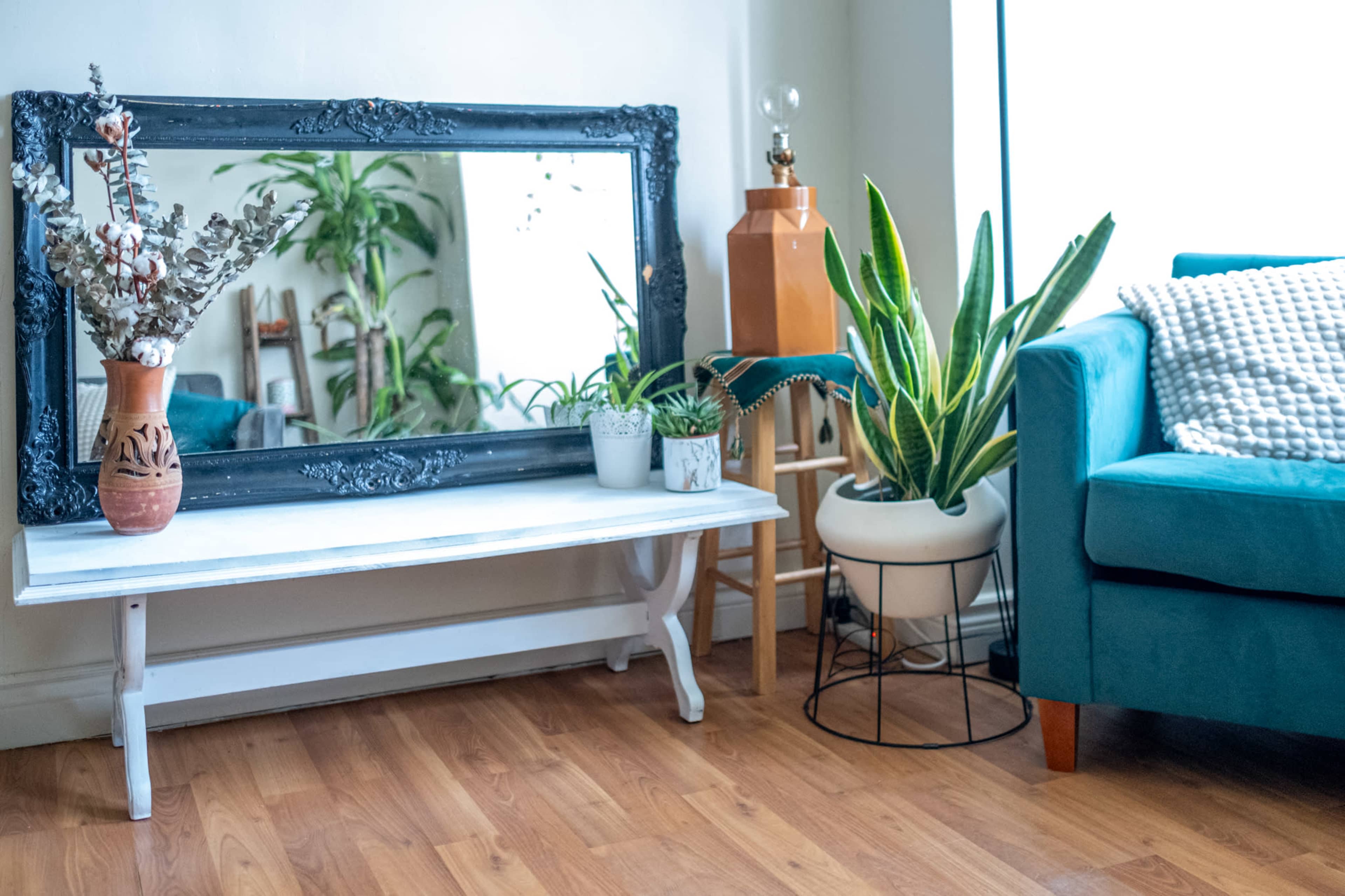 The image shows a cozy living room with a large framed mirror on a white table, surrounded by potted plants and a teal sofa.