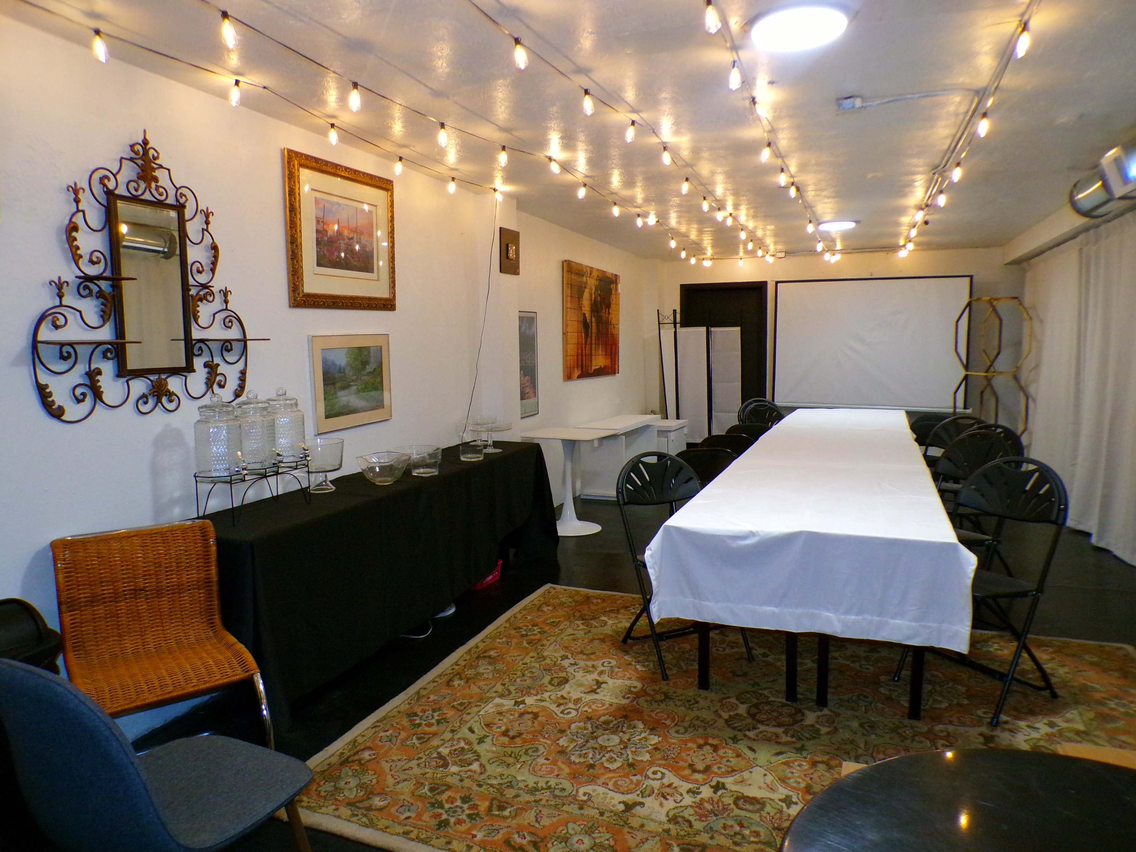 The image shows a well-lit event space with a long table covered in a white tablecloth, surrounded by black chairs, and adorned with string lights overhead.