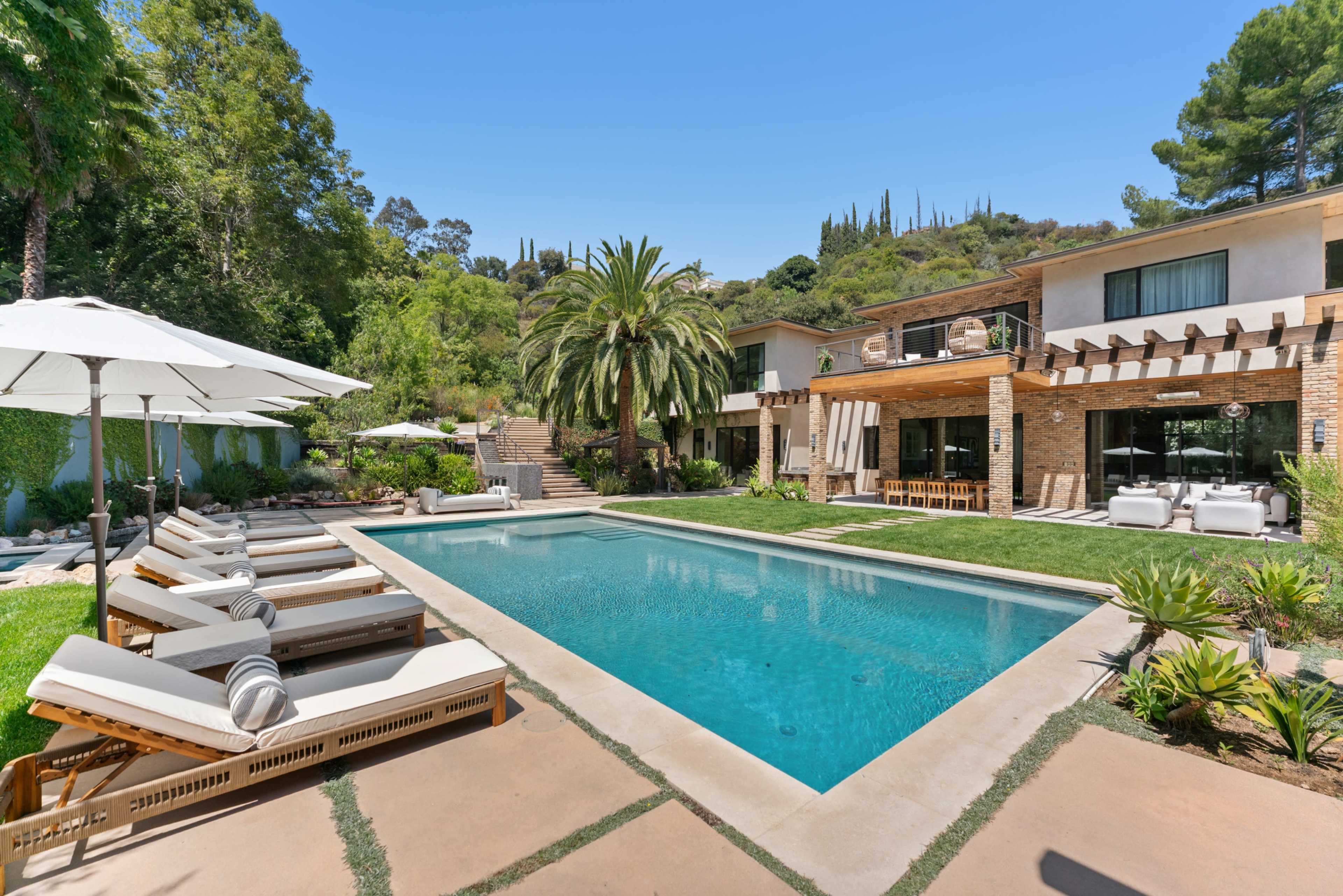 The image features a modern house with a pool surrounded by lounge chairs and palm trees, set in a landscaped garden.