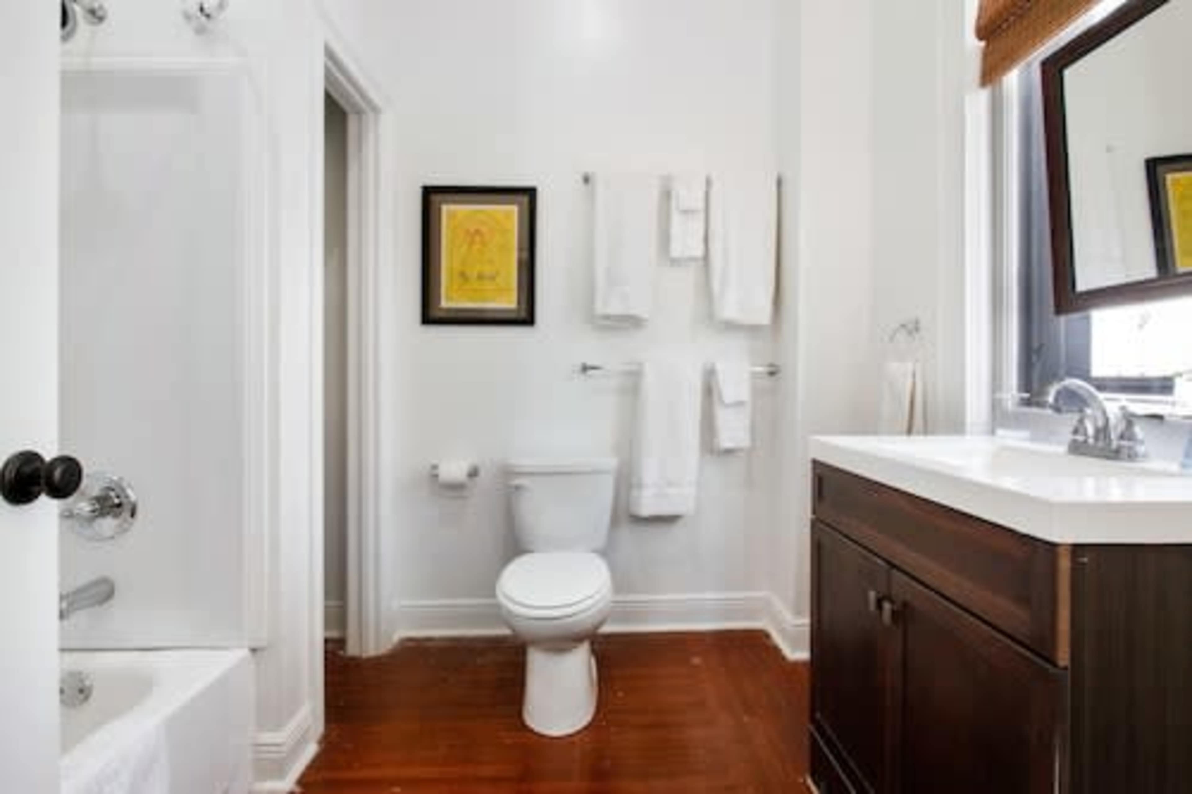 The image shows a clean bathroom with a shower, toilet, sink, and a wooden cabinet, featuring white walls and towels hanging on the racks.