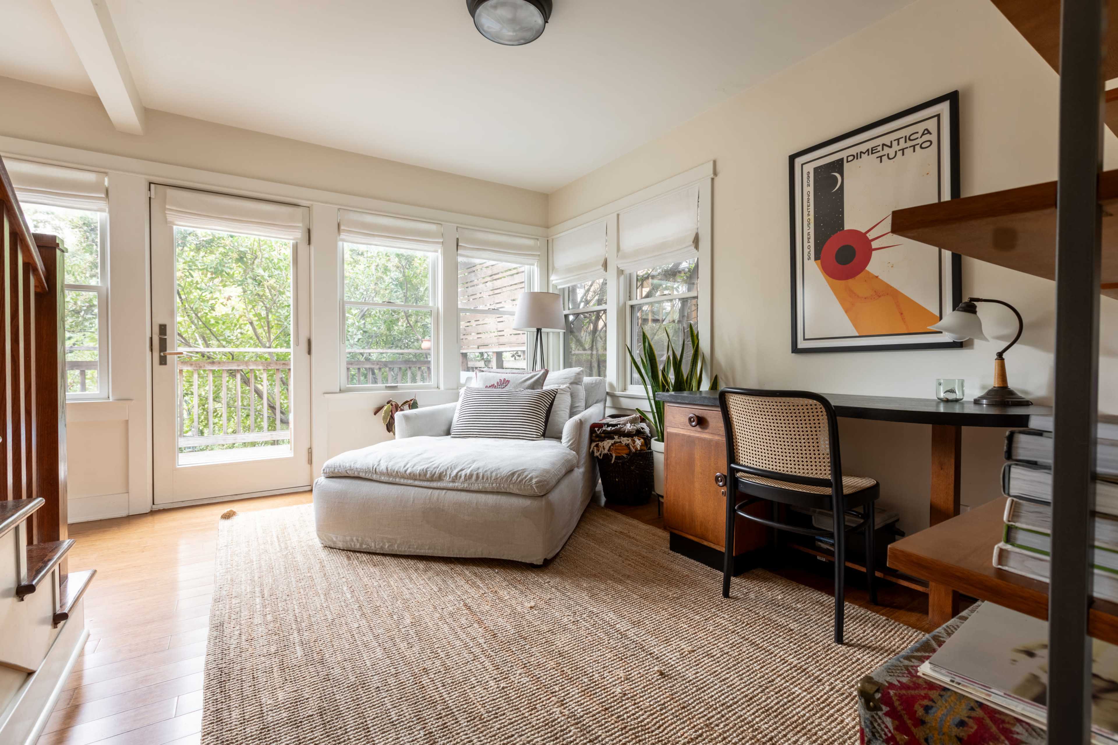 The image shows a cozy room featuring a white chaise lounge, a desk, and large windows allowing natural light, with a woven rug on the wooden floor.