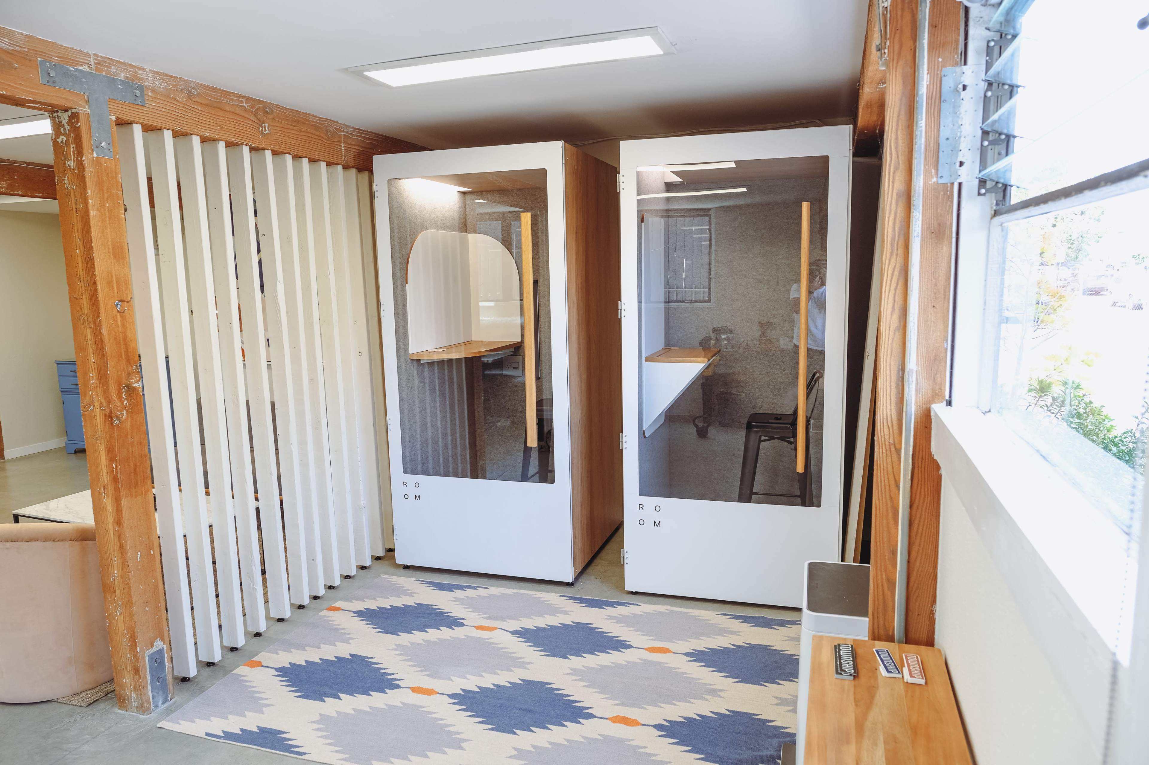 A small, modern workspace with two soundproof booths and a wooden partition, along with a rug and a desk area.