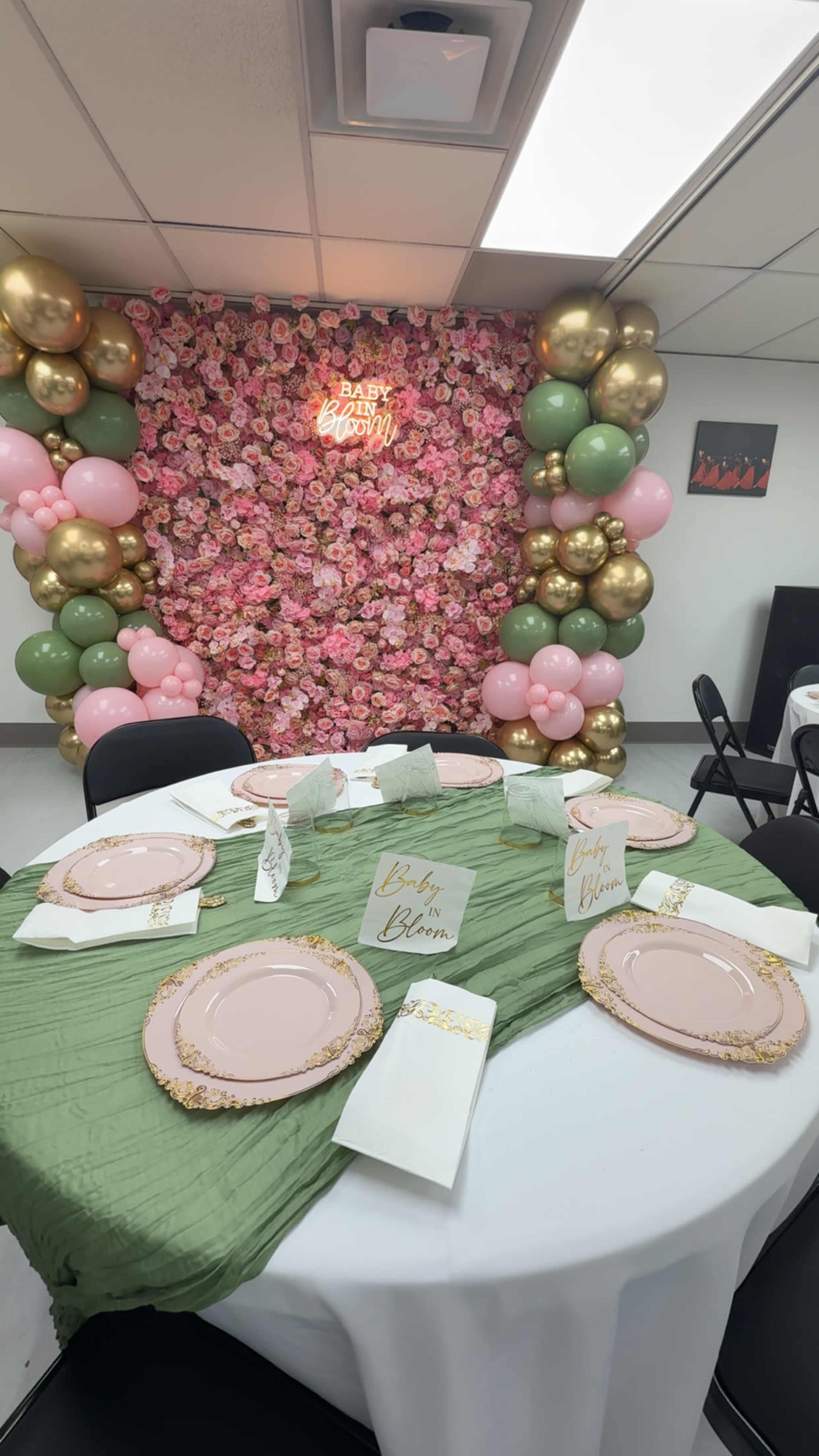 The image shows a decorated event space with a floral backdrop of pink and gold balloons, set with a table featuring green tablecloths, pink plates, and elegant napkins.