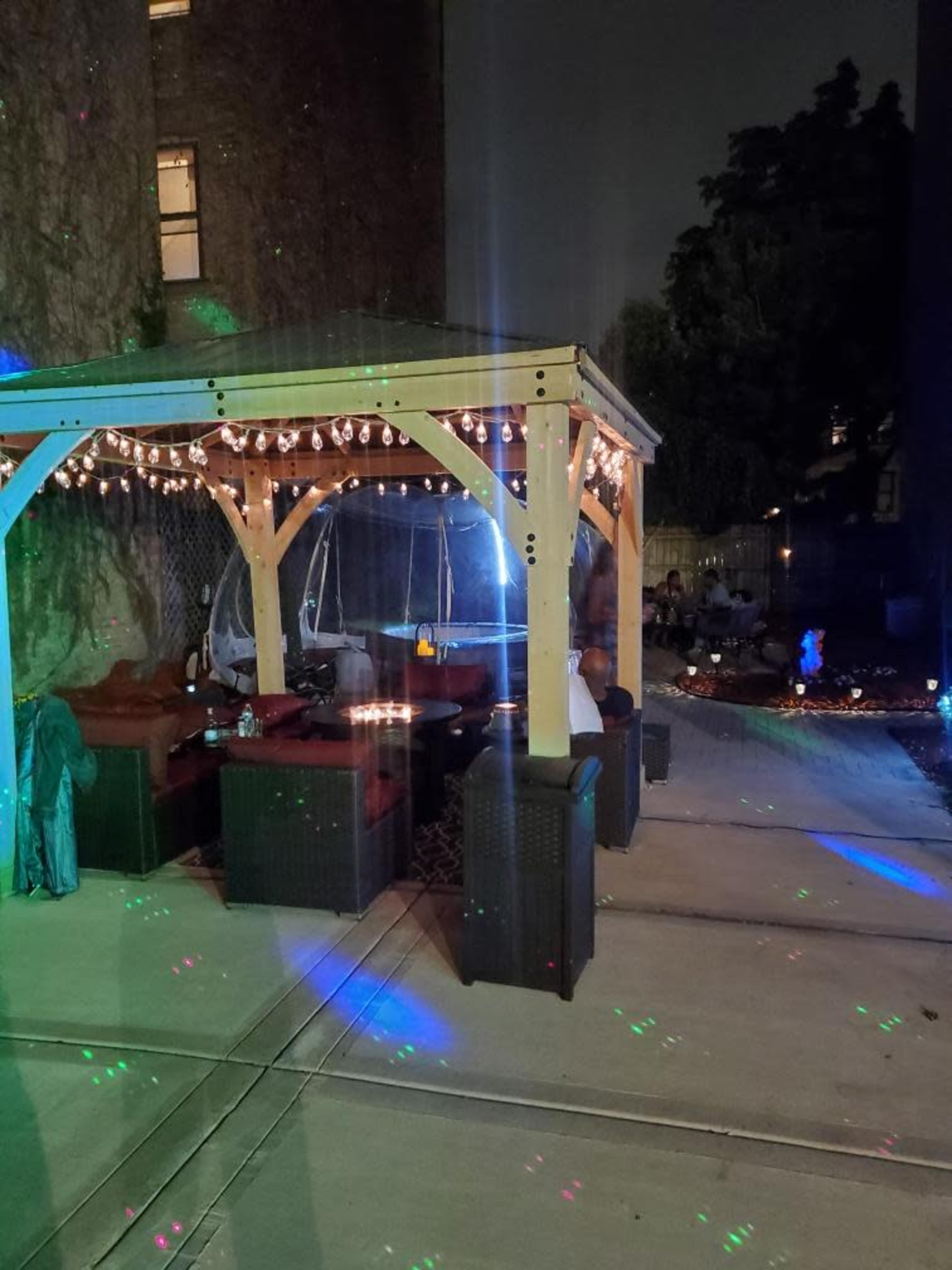 A lit outdoor patio features comfortable seating under a wooden gazebo, with colorful lights reflecting on the ground.