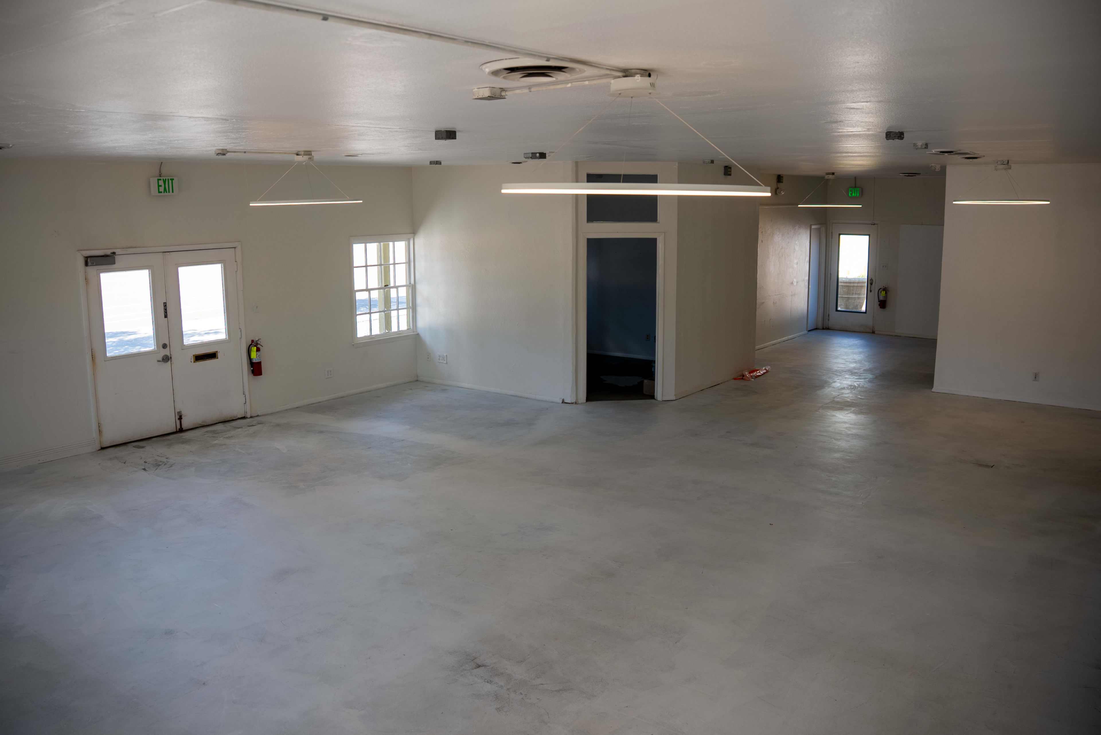 The image shows a spacious, empty room with white walls and a concrete floor, featuring two entrances and overhead lighting.