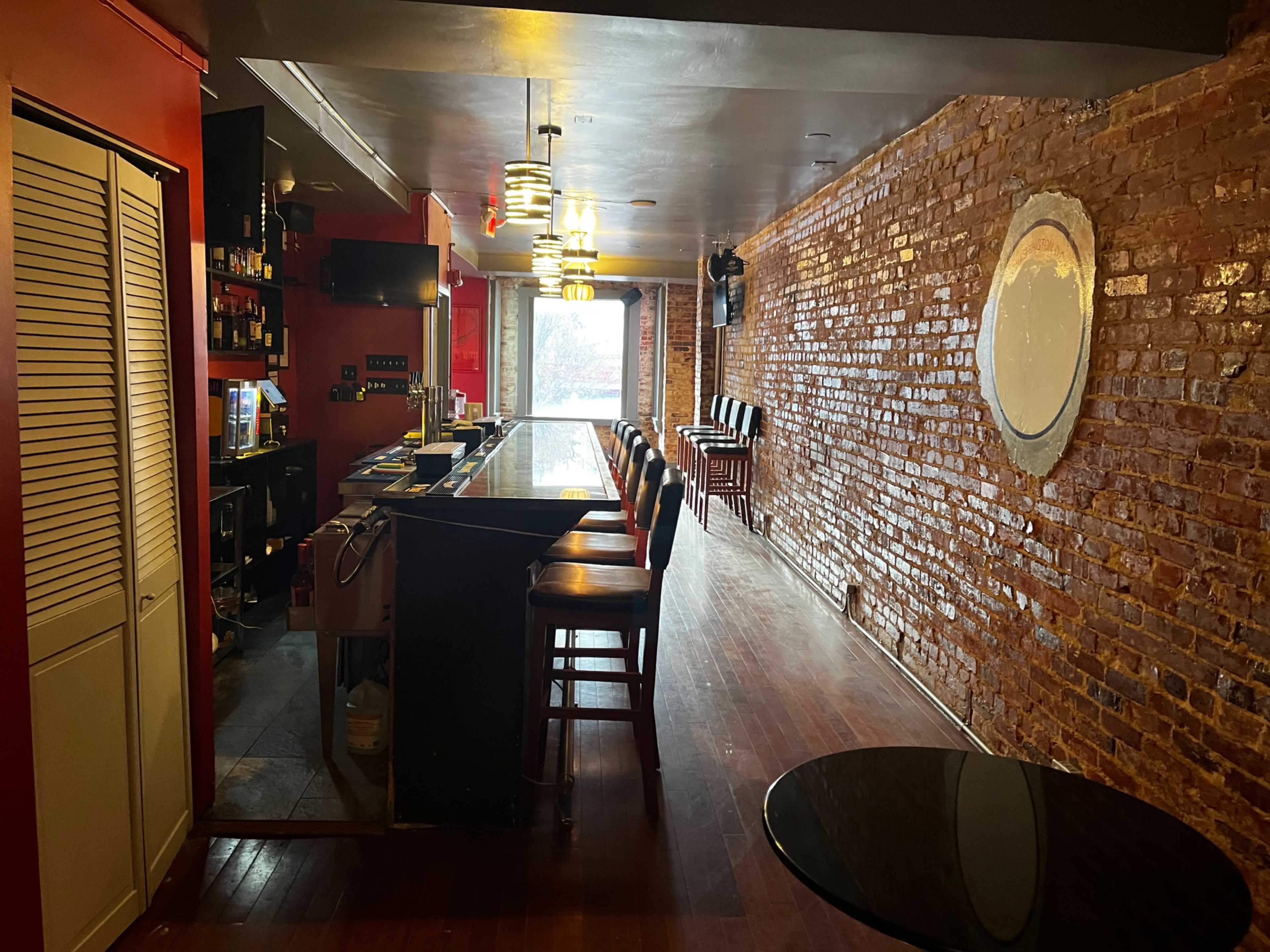 An empty bar with a polished counter and exposed brick walls, featuring a few chairs along one side and a window at the far end.