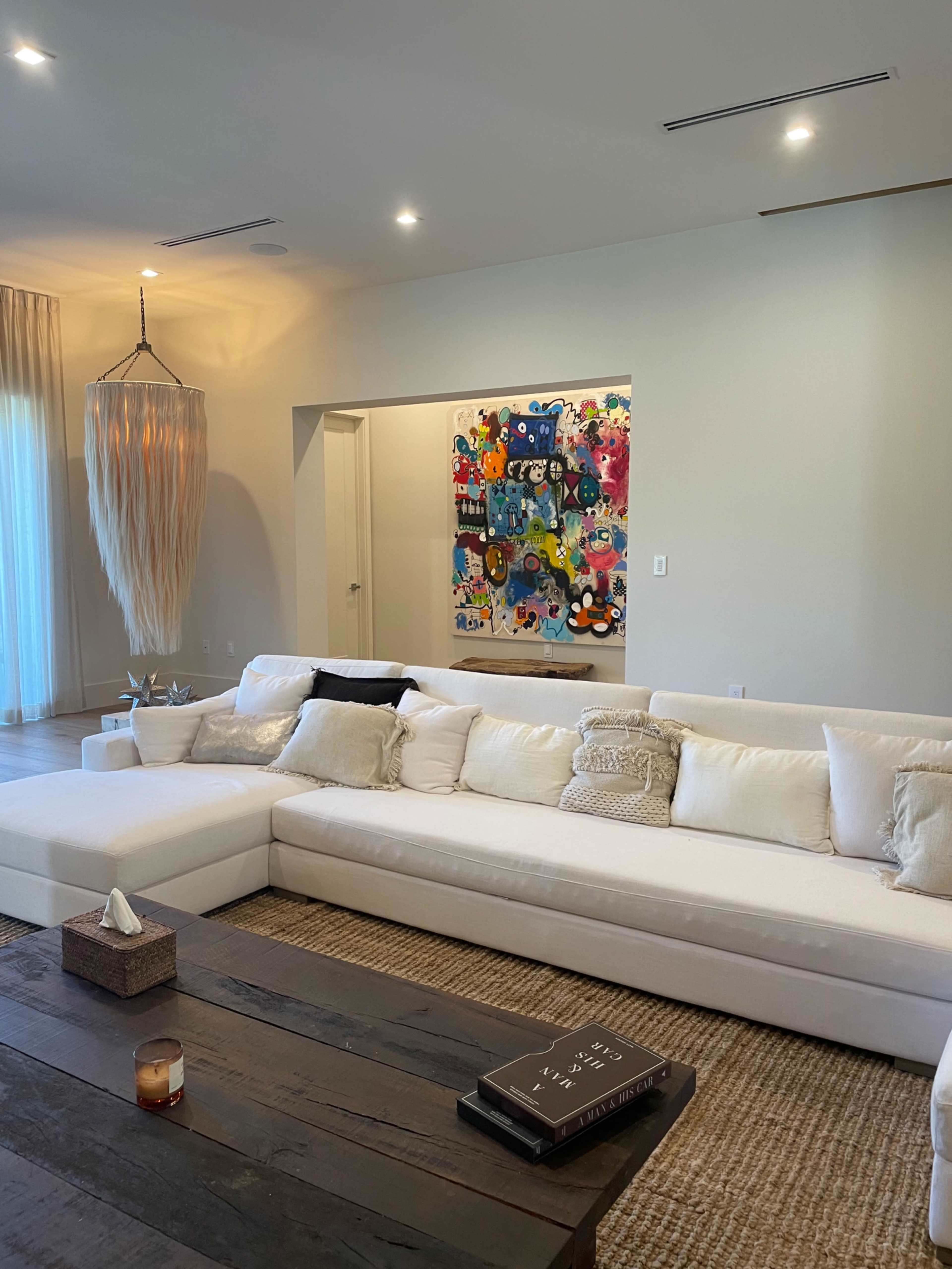 The image shows a modern living room featuring a large white sectional sofa, a wooden coffee table with a book, and a colorful abstract painting on the wall.