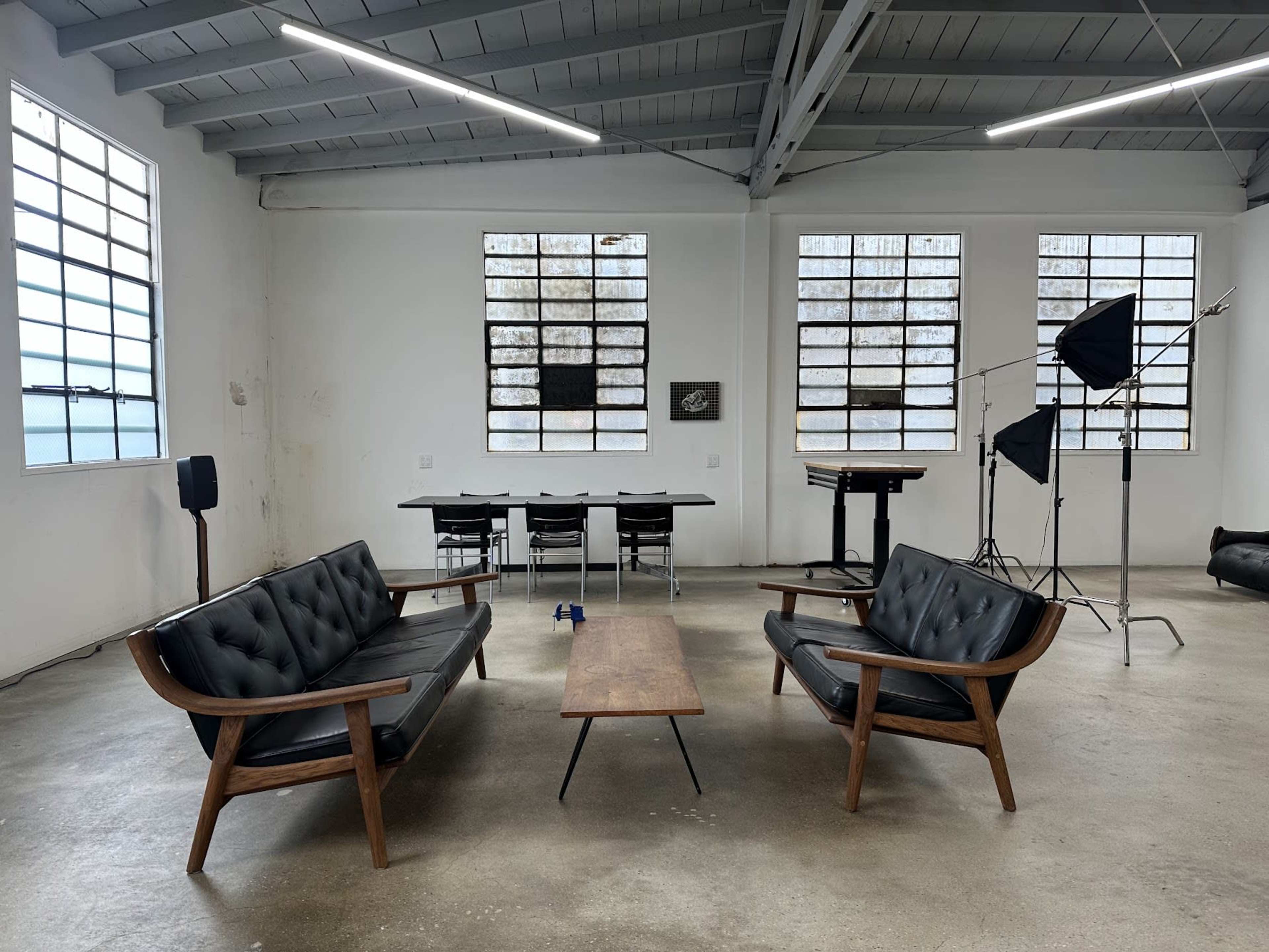 A minimalist interior featuring two black leather sofas facing a wooden coffee table, with industrial-style windows and a backdrop of studio equipment.