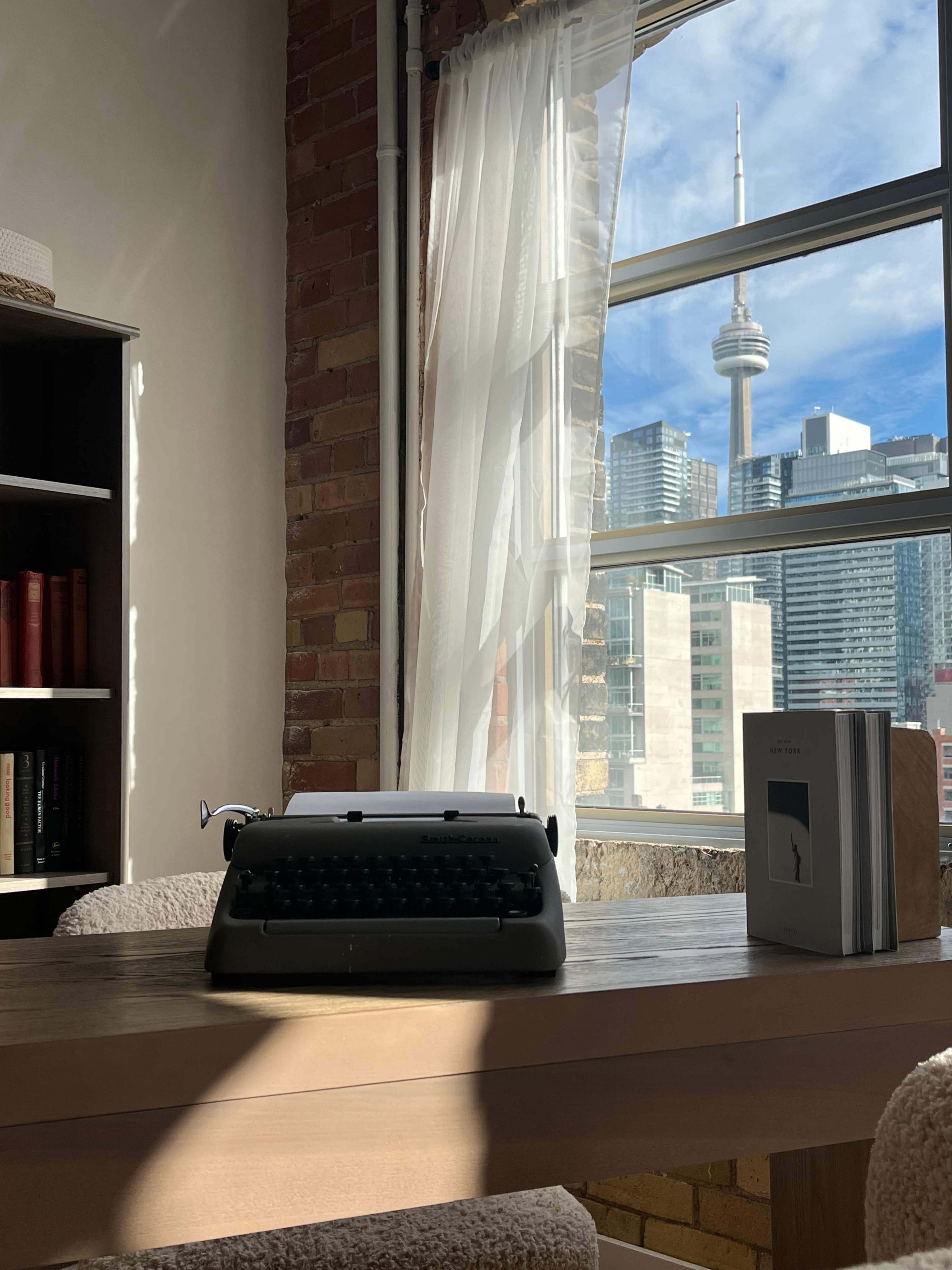 A vintage typewriter sits on a wooden desk beside a window showcasing a view of the CN Tower and a city skyline.