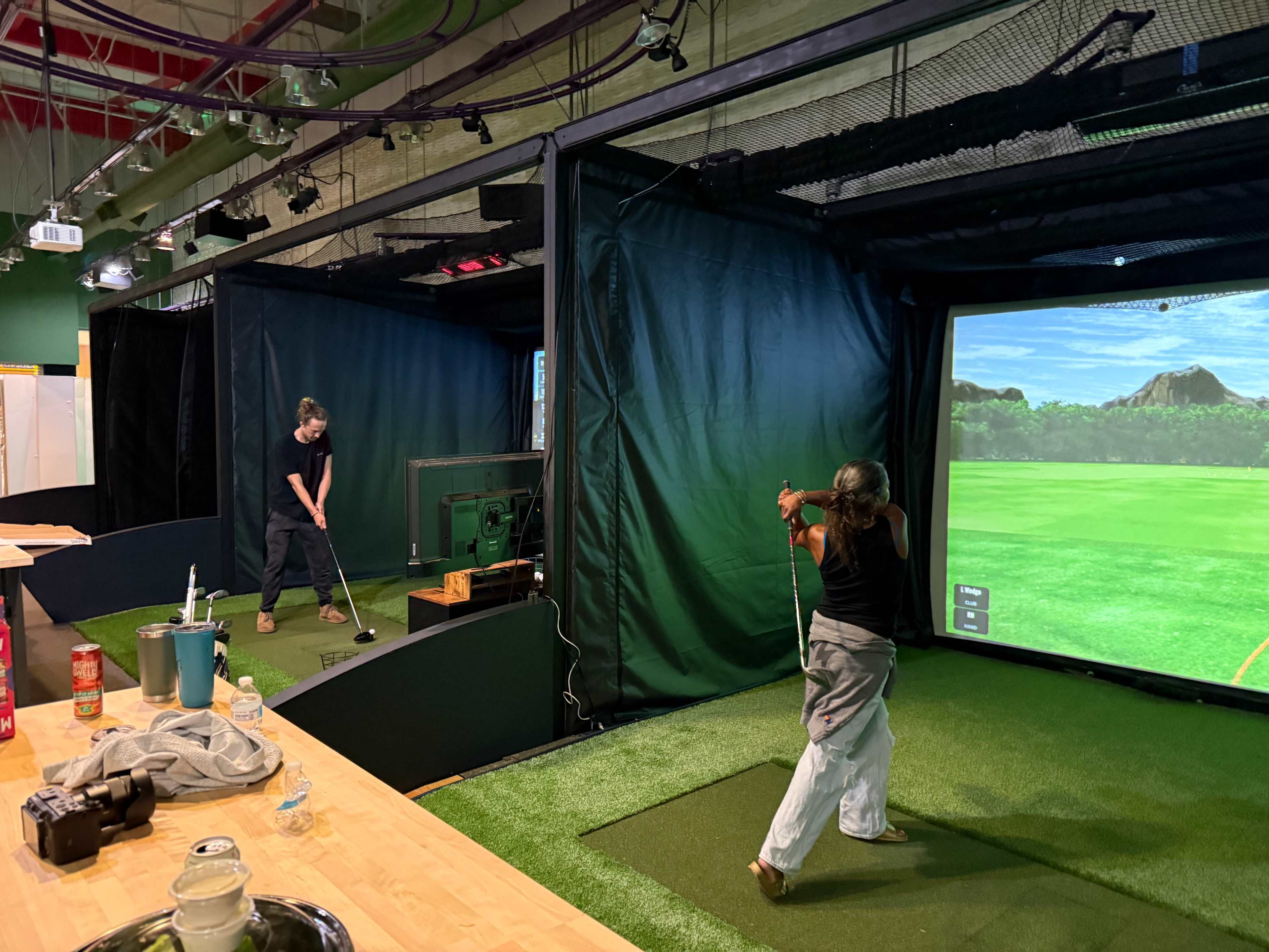 Two individuals practice golf swings in a simulated indoor golf environment, with a large screen displaying a virtual course in the background.