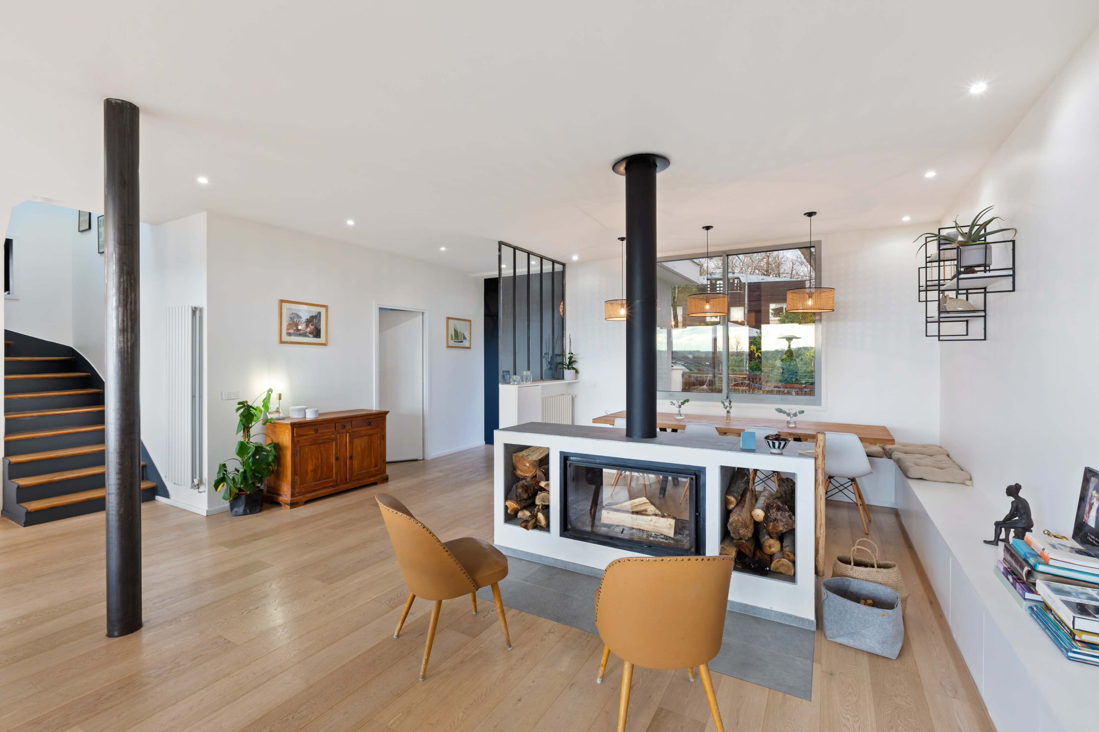 The image shows a contemporary living space featuring a centrally located fireplace, a dining area with a long table, and a staircase leading to an upper level.