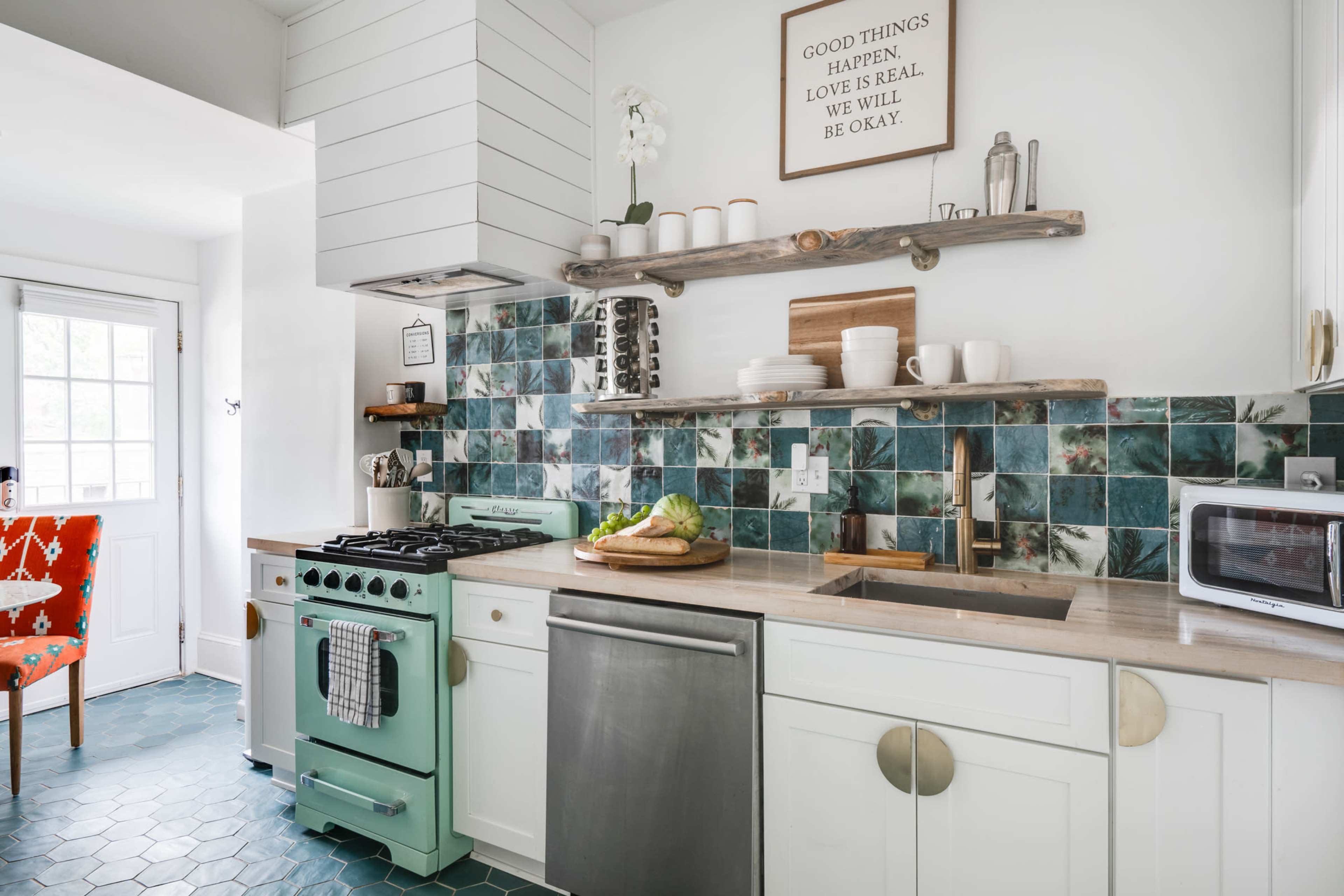 The kitchen features a mint-green stove, blue-green backsplash tiles, and a wooden shelf holding decorative items, with a view of a door leading outside.