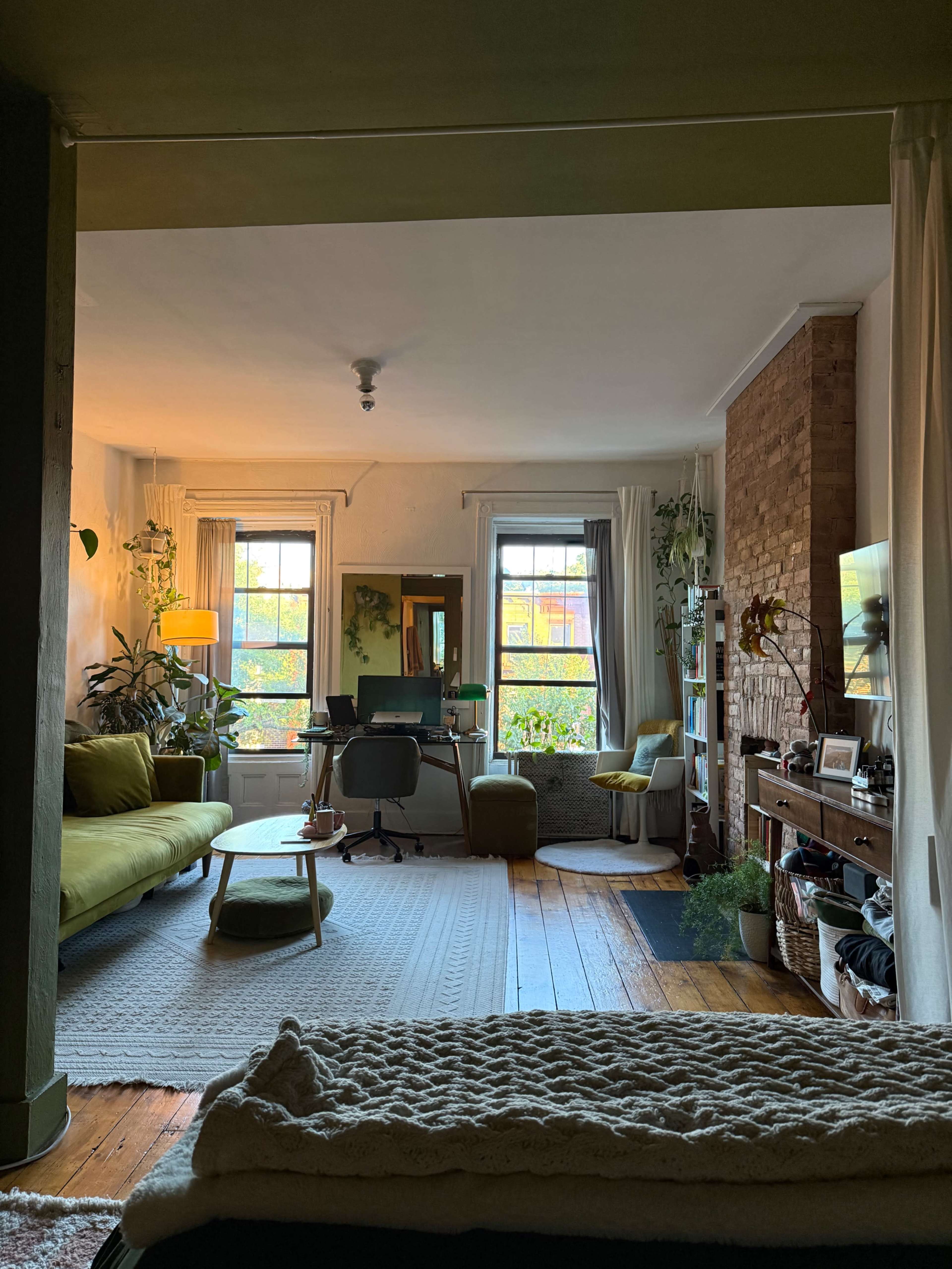 The image shows a cozy living room with a green sofa, a desk, and large windows letting in natural light, surrounded by plants and wooden flooring.