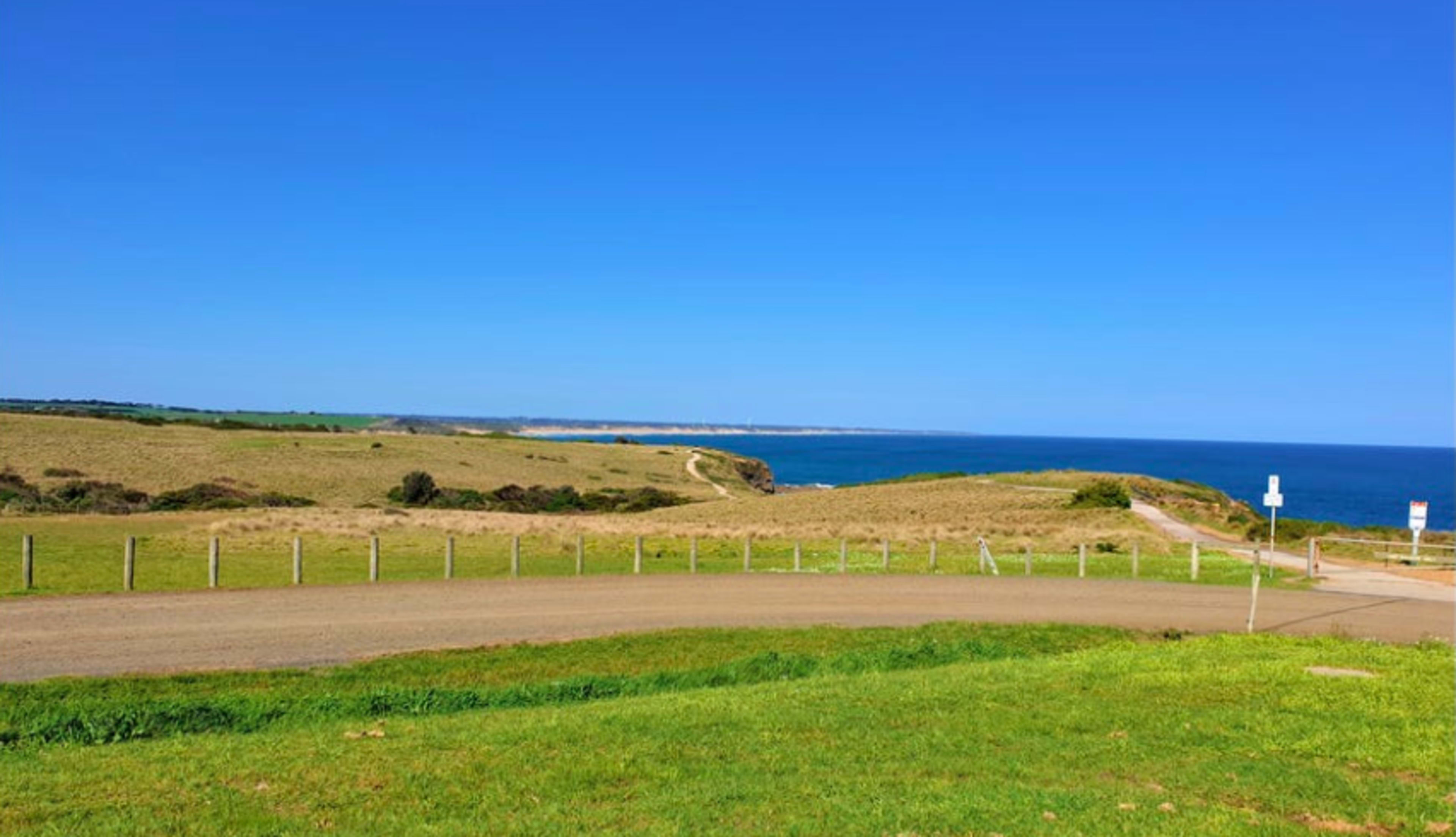 The image shows a coastal landscape with grassy fields leading down to the ocean under a clear blue sky.