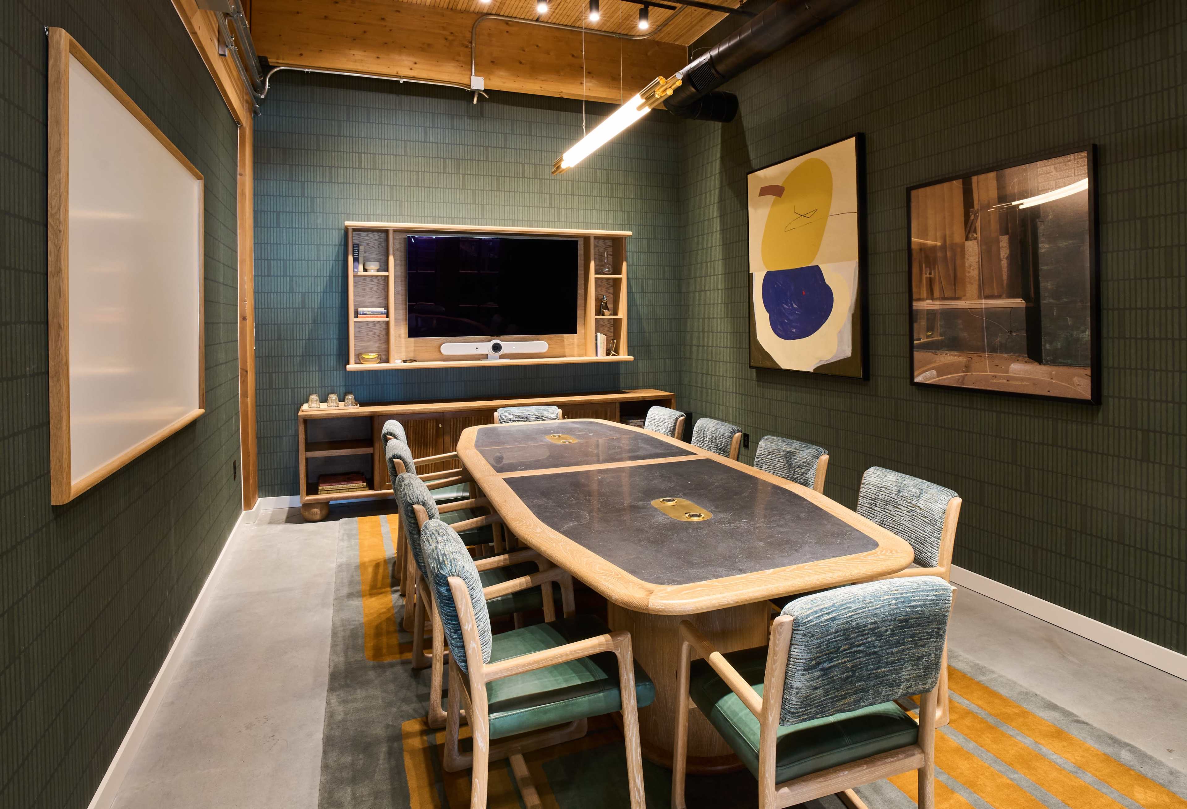 A conference room with a large oval table surrounded by eight chairs, green walls, and a display screen mounted on the back wall.