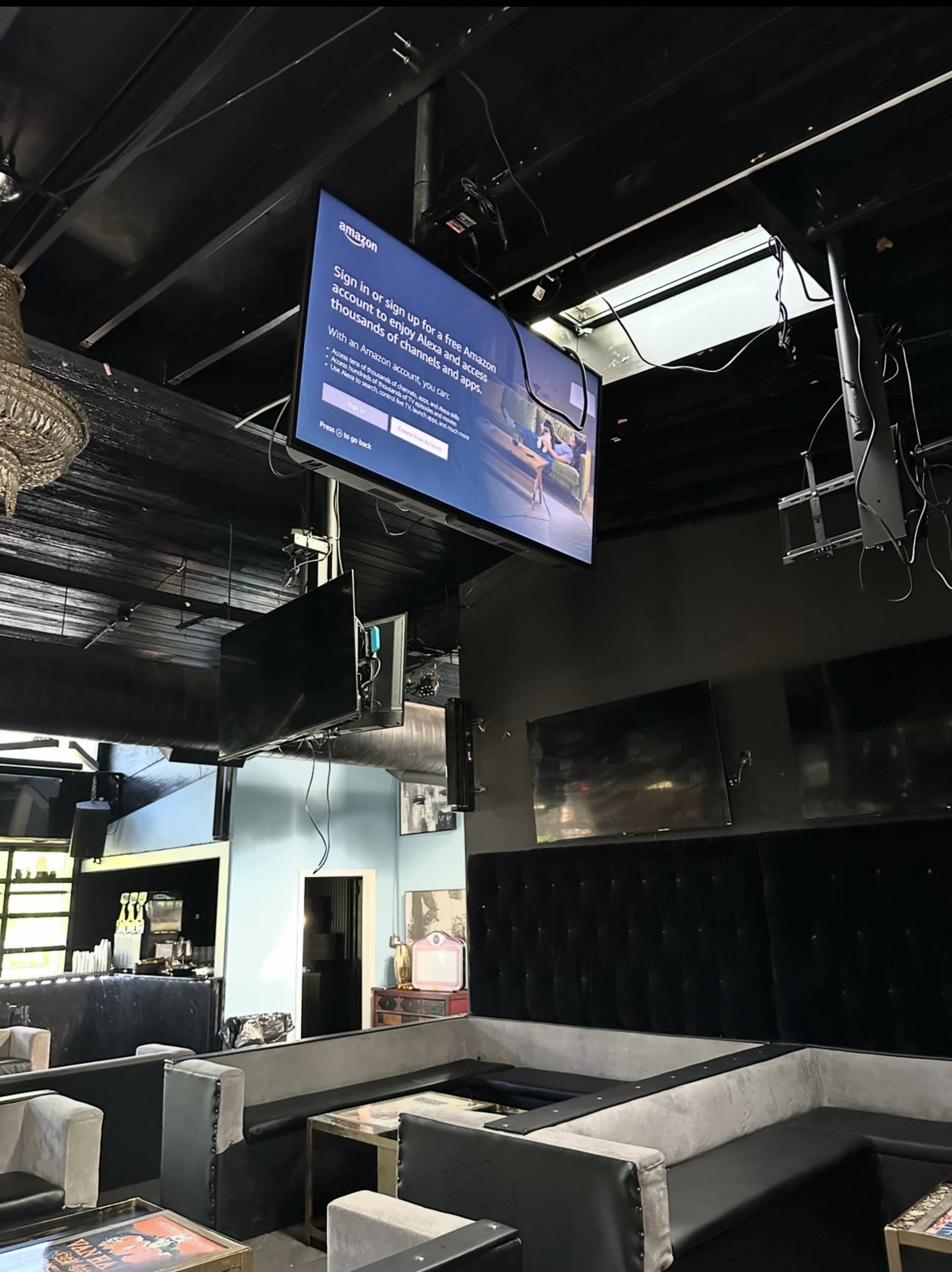 The image shows the interior of a bar or lounge with a large television mounted on the ceiling displaying an Amazon login screen, surrounded by dark decor and seating.