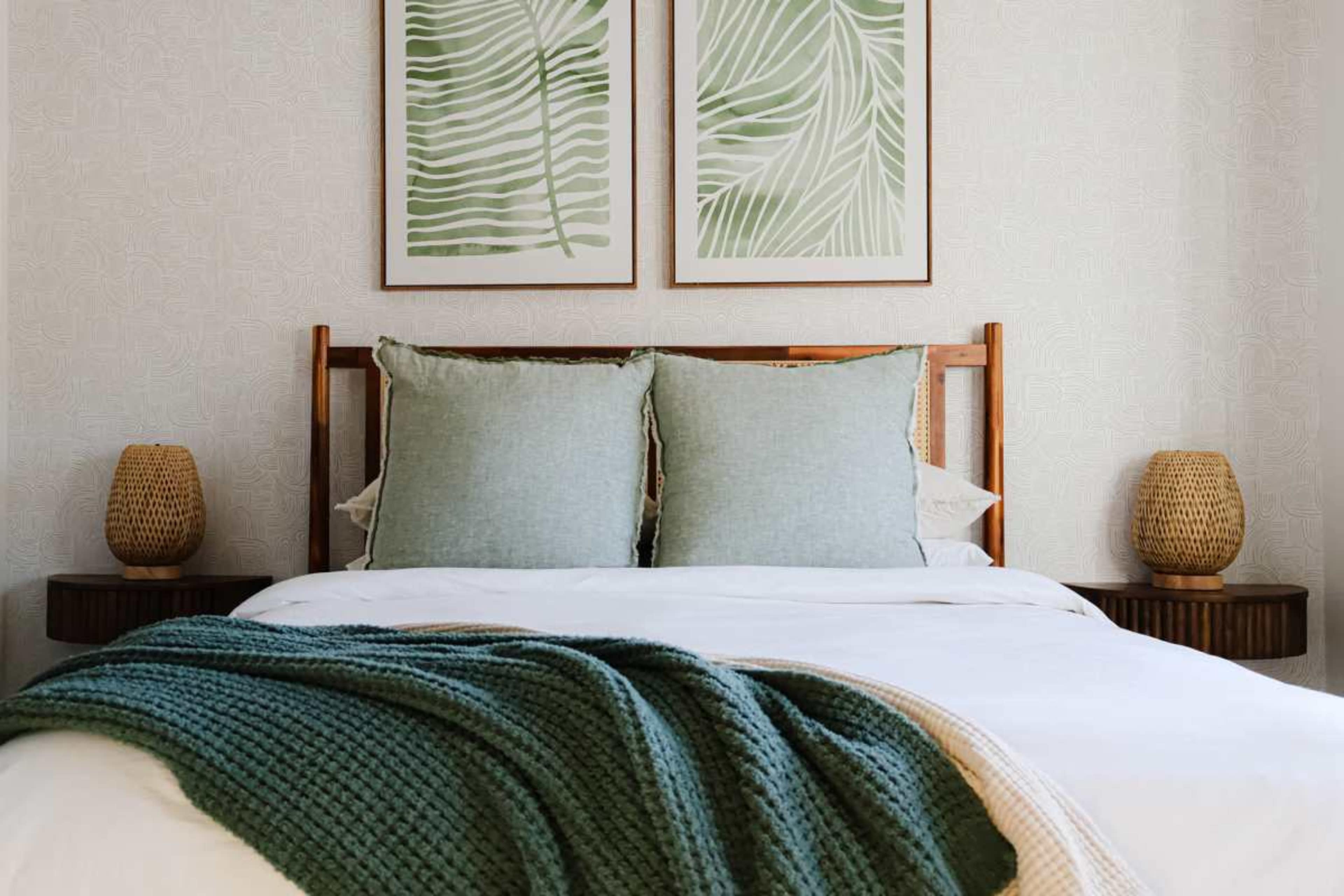 A bed with decorative pillows and a green throw blanket is flanked by two woven lamp stands and features framed botanical prints on the wall.