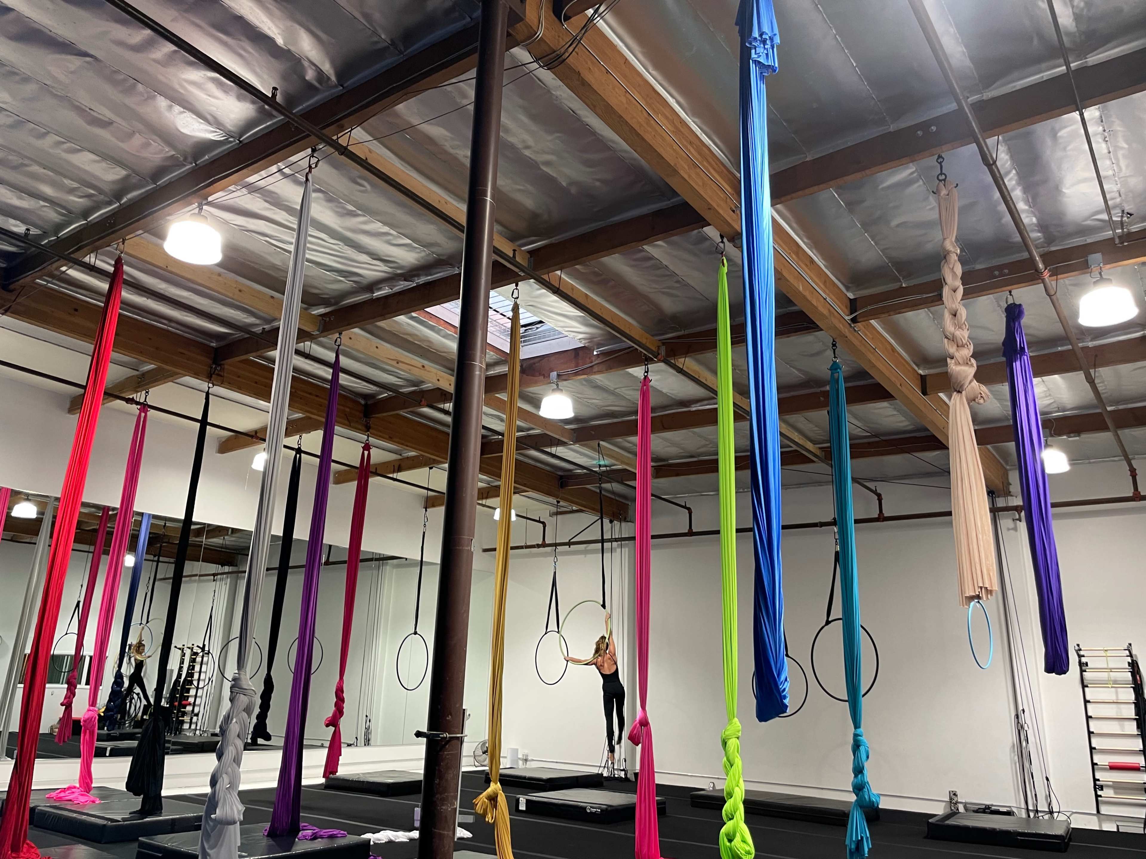 A gym studio with multiple hanging silk aerials in various colors and a person practicing on a suspended hoop.