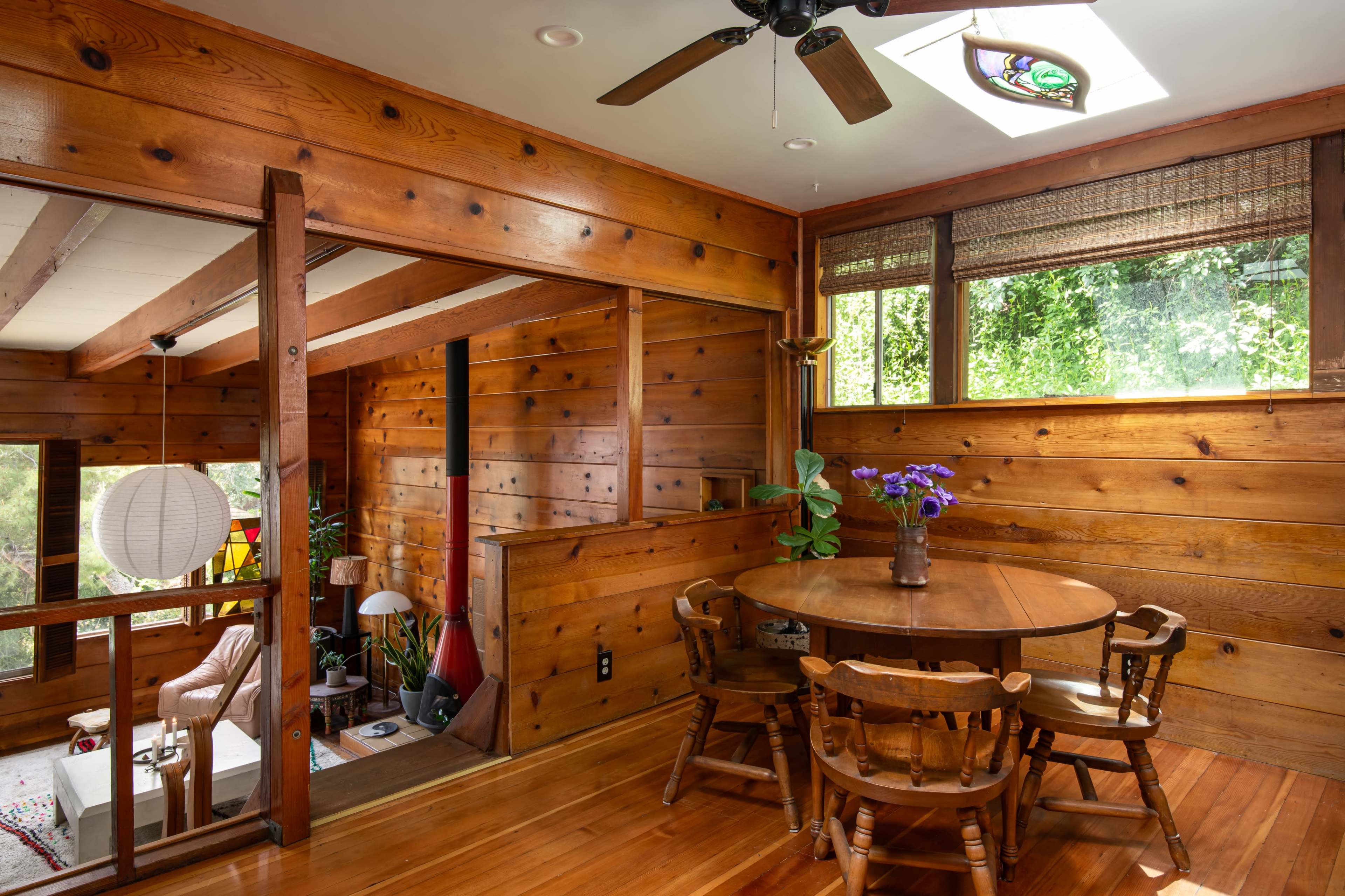 The image shows a wooden interior of a home with a round dining table, four chairs, and large windows letting in natural light.