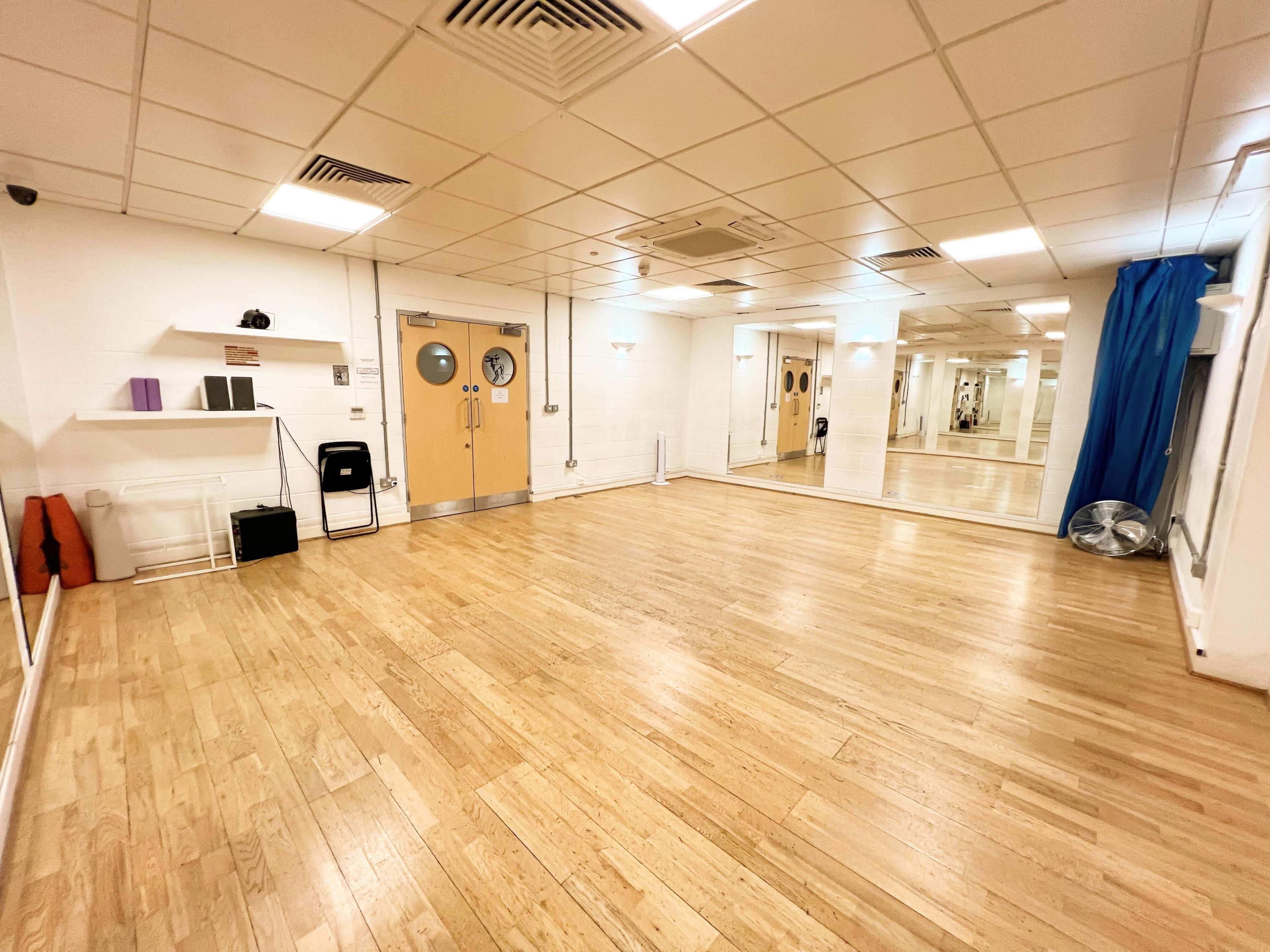 The image shows an empty fitness studio with wooden flooring, mirrors on one wall, and large doors with round windows.