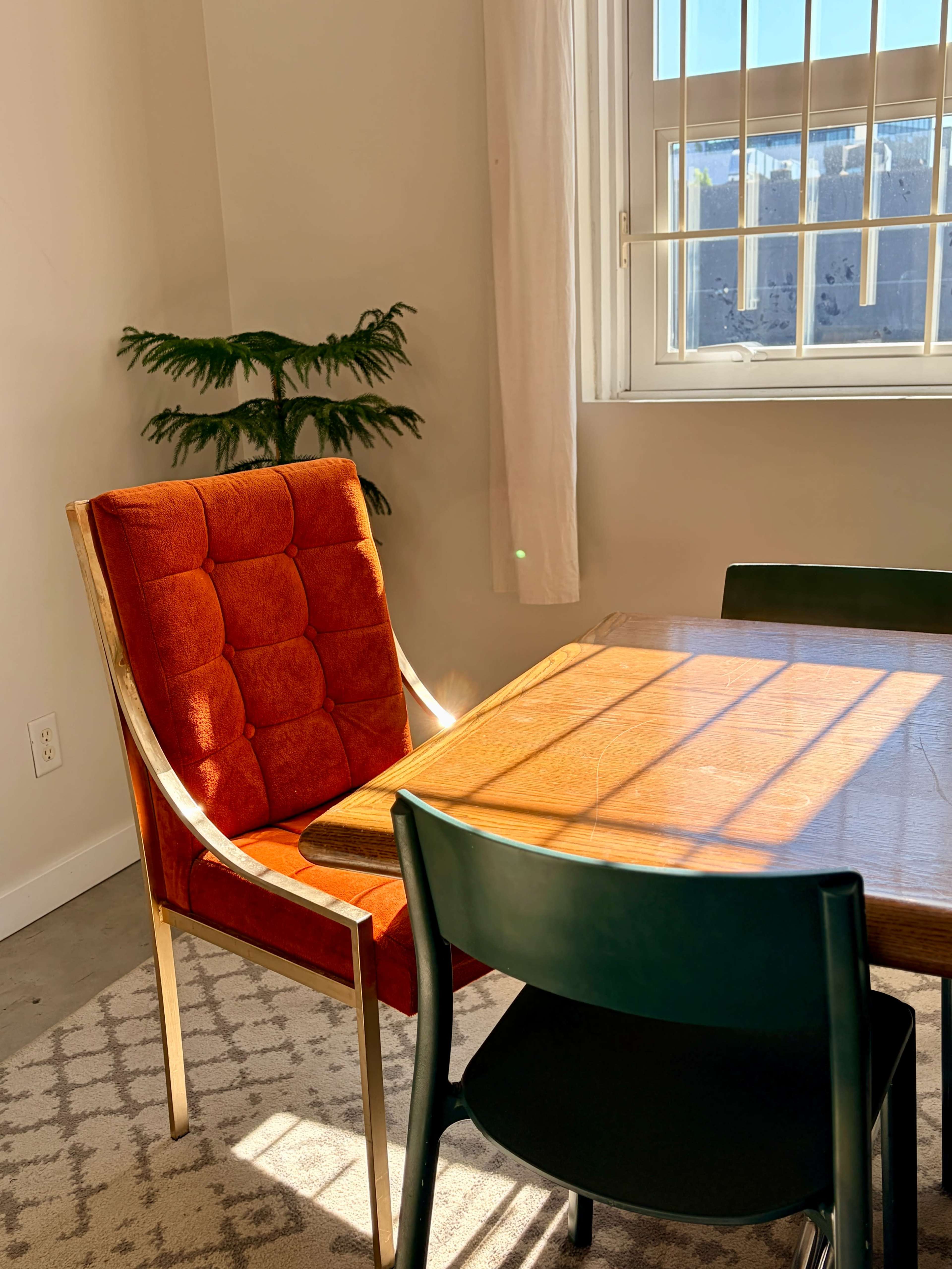 A wooden table with a red upholstered chair and a green chair sits in a well-lit room beside a window with sunlight casting shadows on the floor.