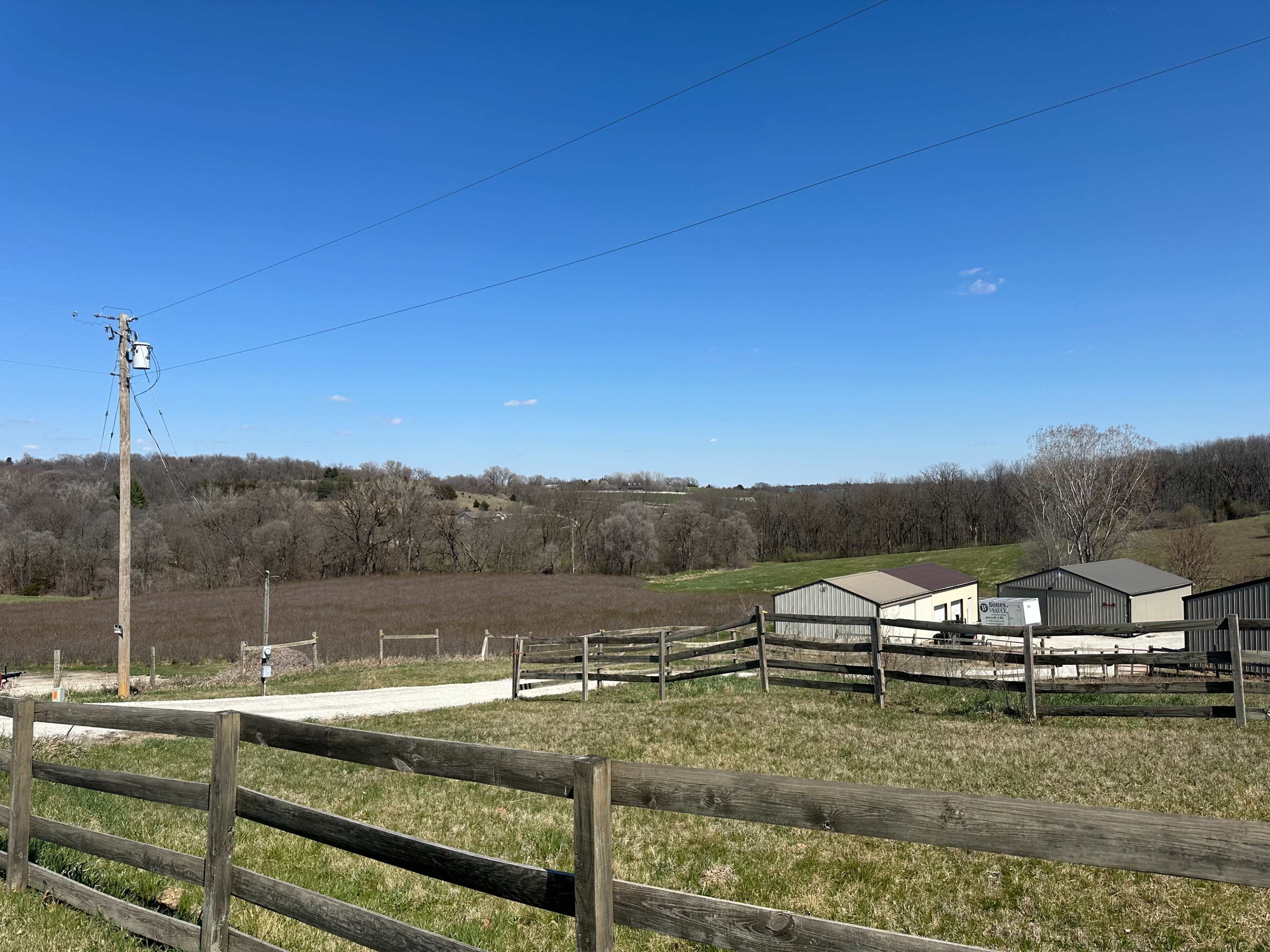 A rural landscape features a clear blue sky, several farm buildings, a fence line, and rolling hills in the background.