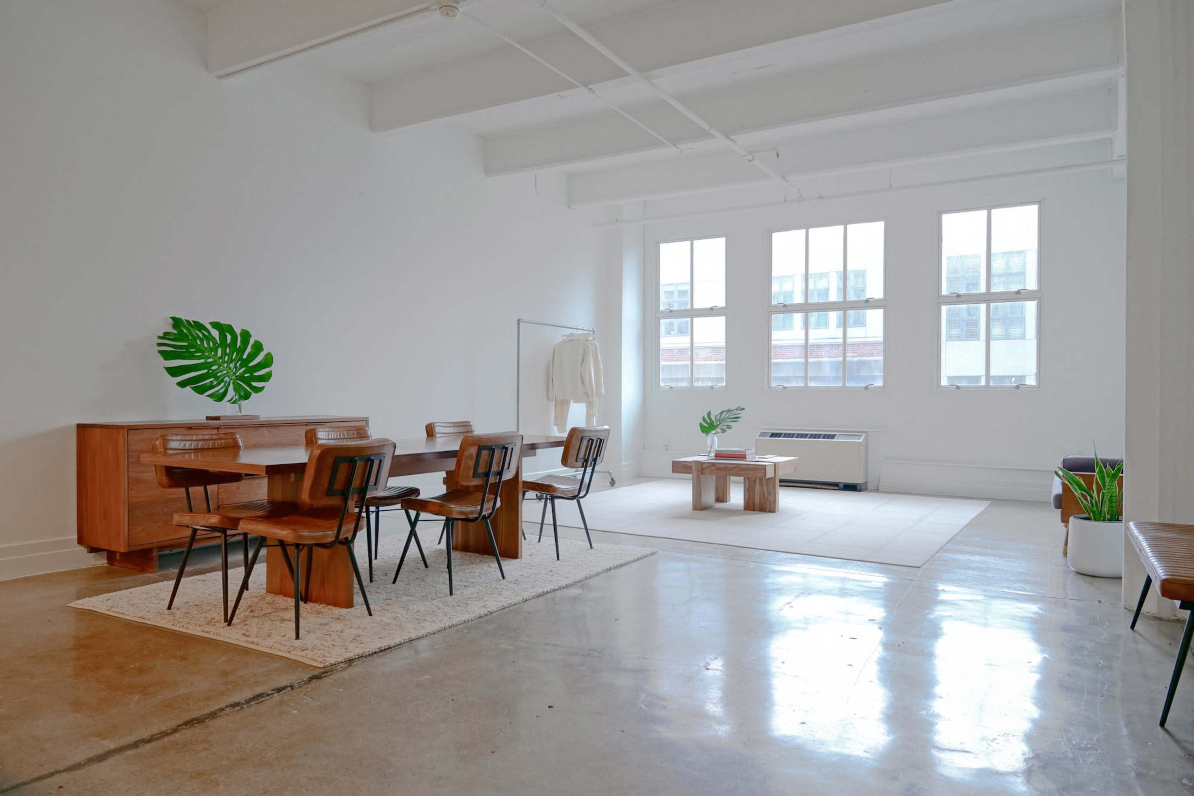 A bright, minimalist interior space featuring a dining area with a wooden table and chairs, and a separate seating area with a small coffee table and plants.