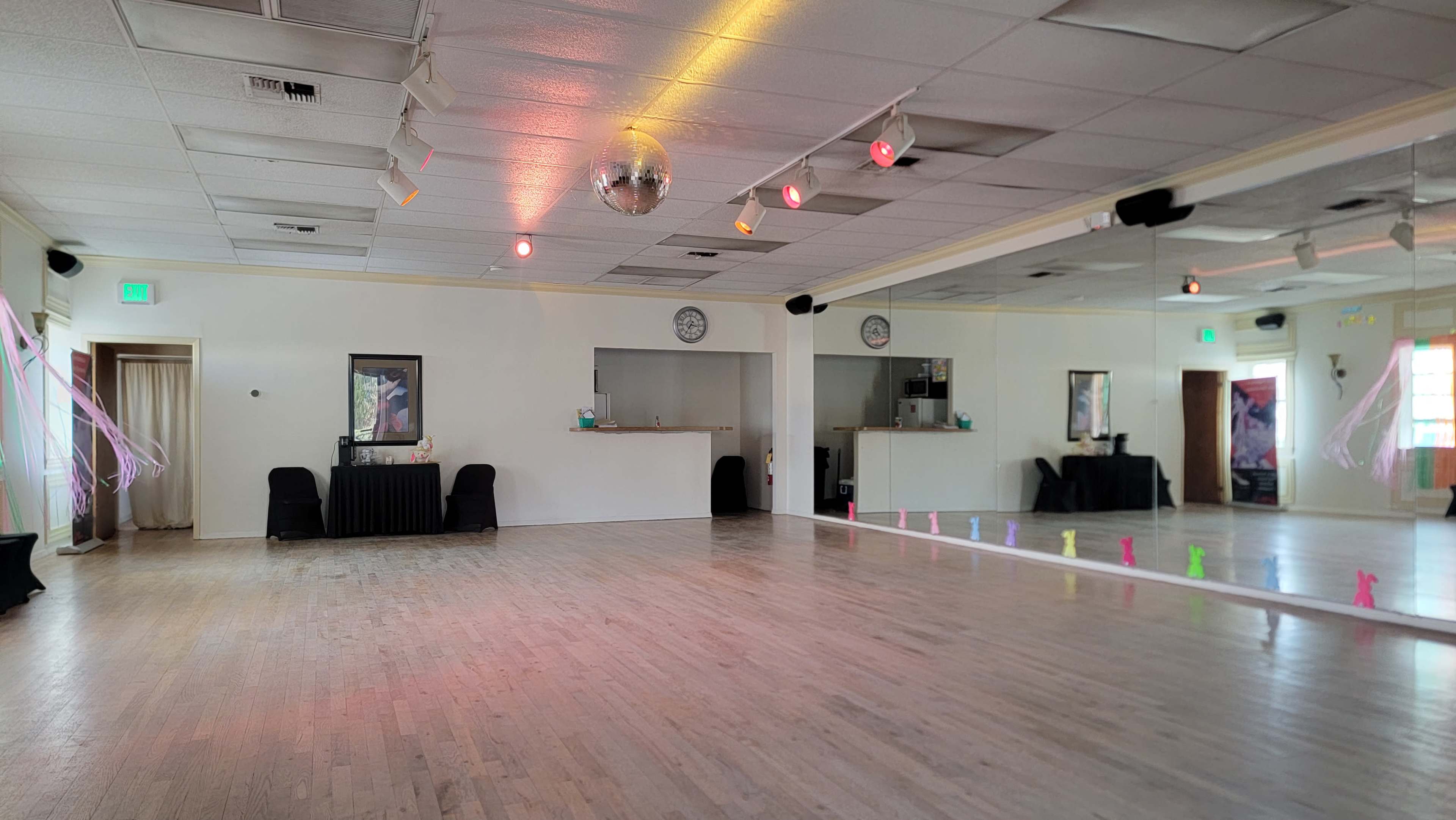 The image shows a spacious dance studio with a wooden floor, wall mirrors, and colored lighting fixtures, featuring a disco ball hanging from the ceiling.