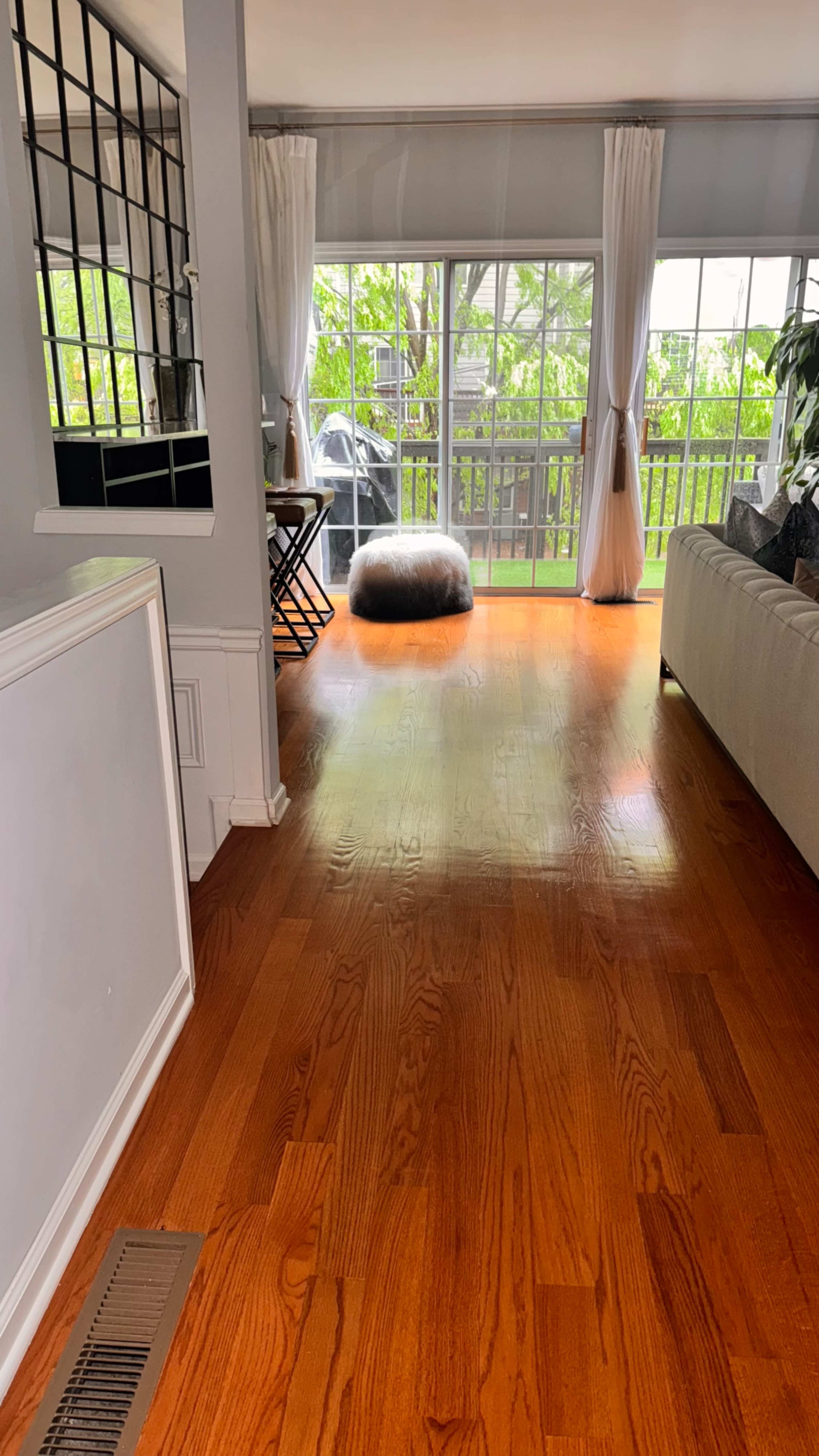 The image shows a room with hardwood floors, a light-colored sofa, and large windows letting in natural light, leading to an outdoor view.