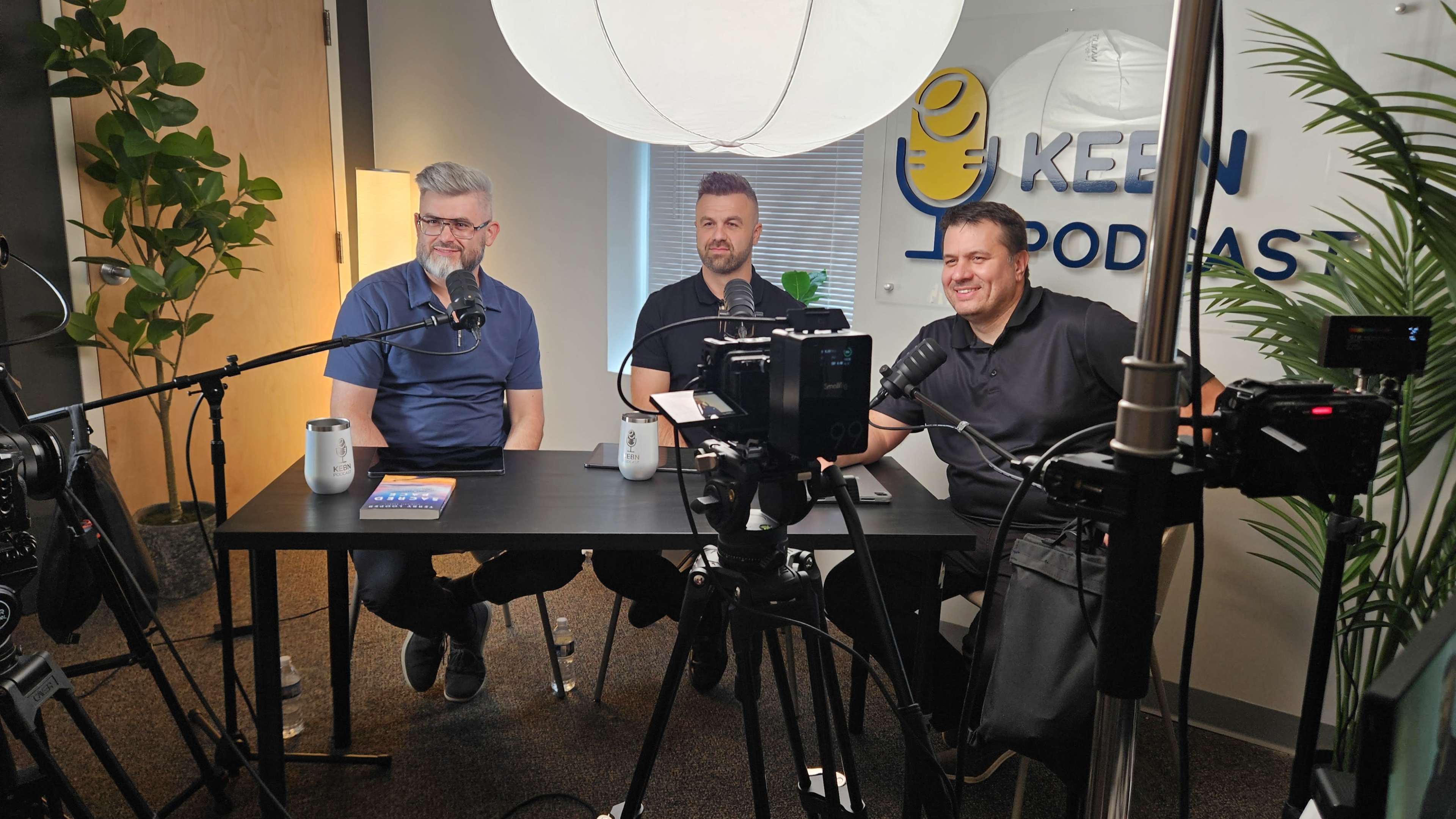 Three men sit at a table in a podcast studio, with microphones and cameras set up around them, ready to record a session.