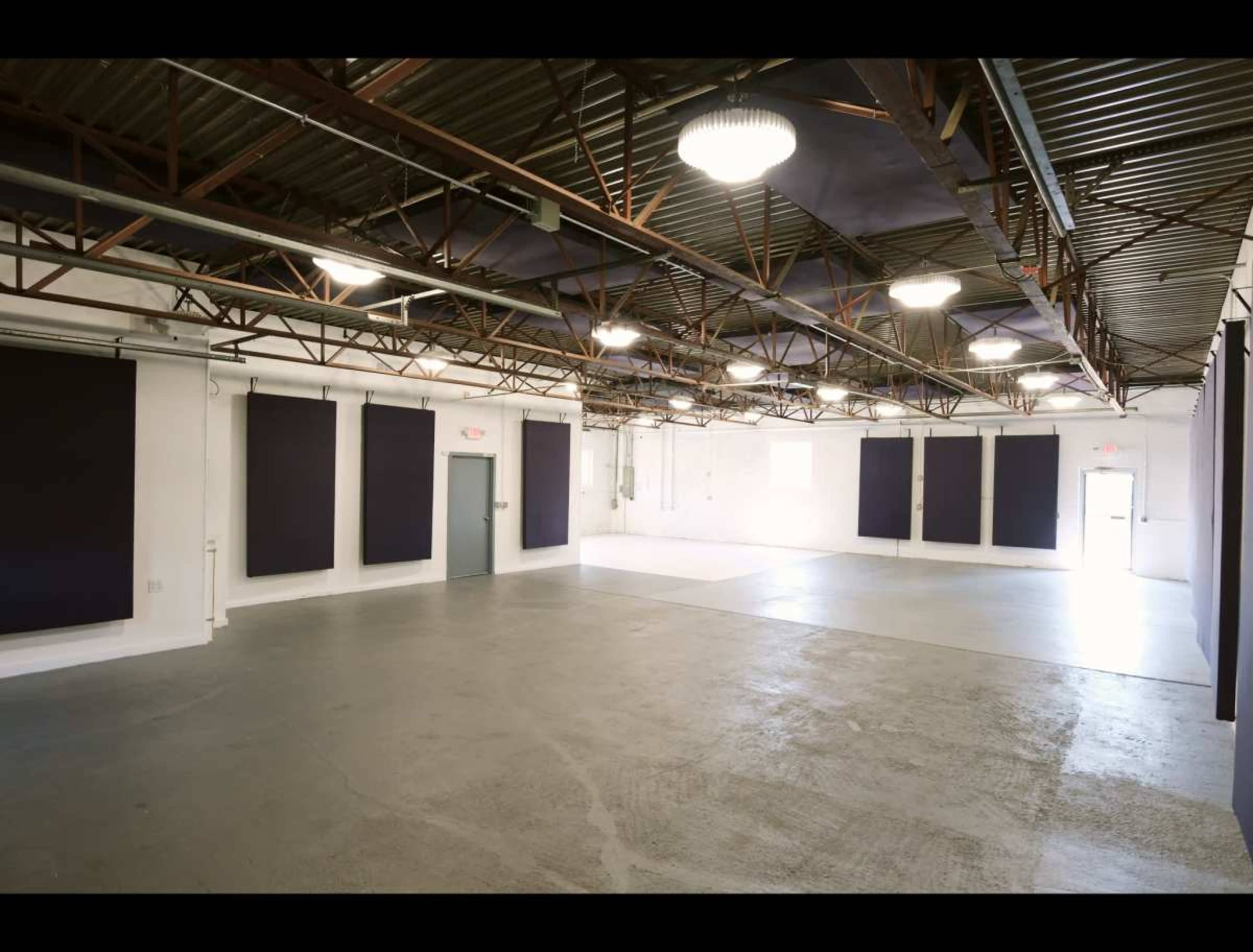The image shows an empty industrial space with concrete flooring and white walls, featuring dark wall panels and overhead lighting.