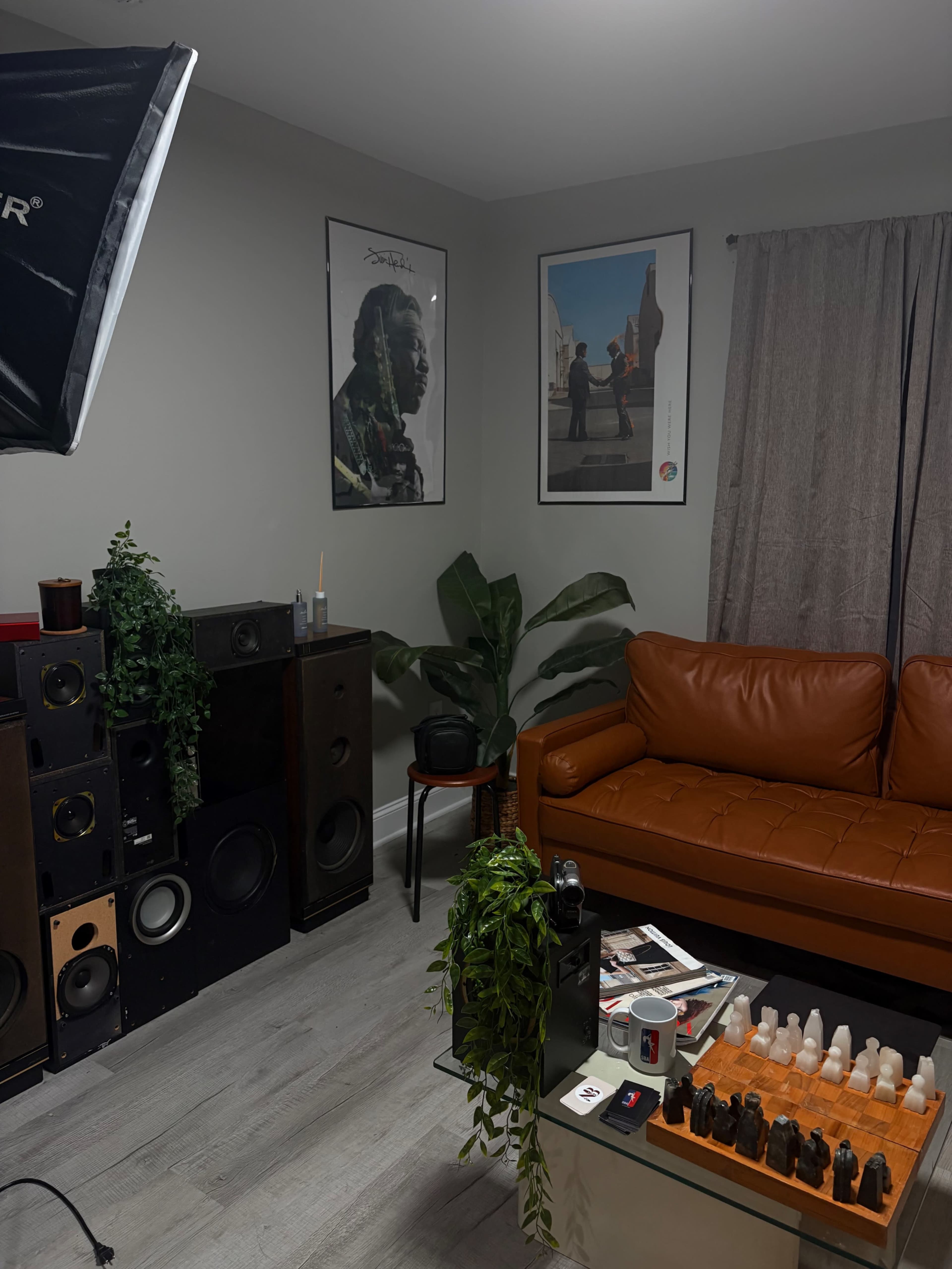 The image shows a cozy living room with a brown leather sofa, a coffee table displaying various bottles, large speakers against one wall, and two framed posters hanging above the sofa.