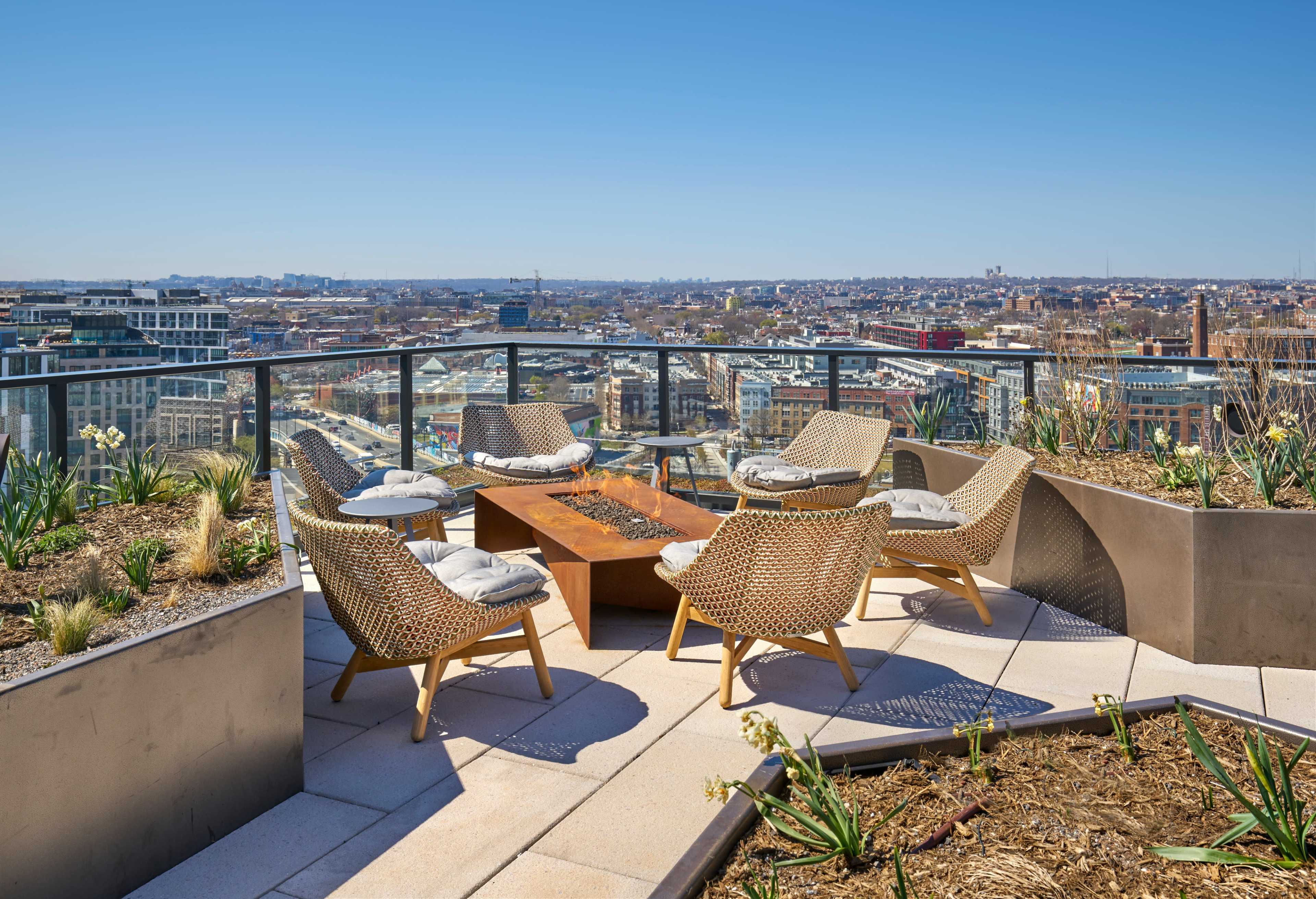 A rooftop terrace features a seating area with wicker chairs around a fire pit, overlooking a city skyline on a clear day.