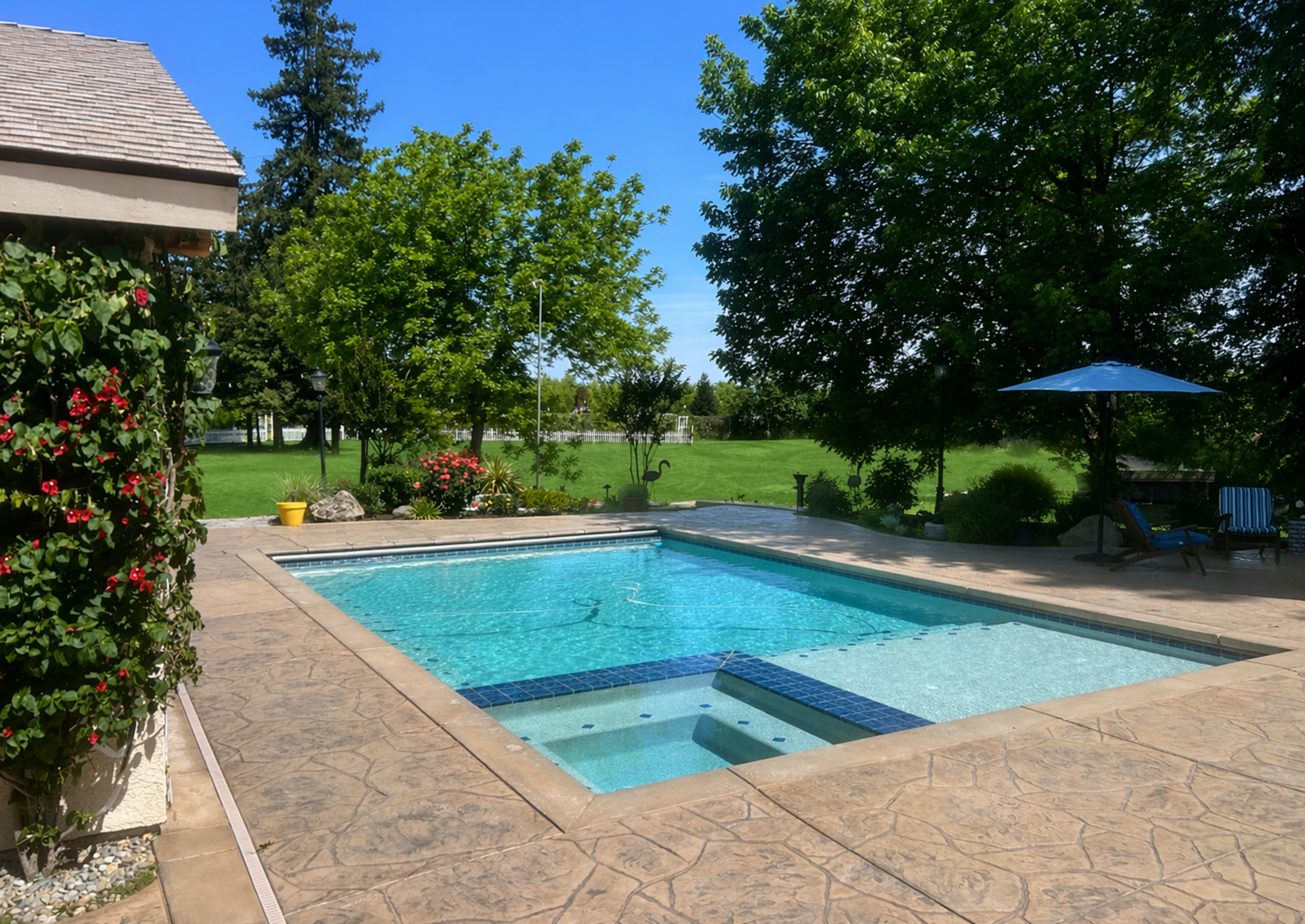 The image shows a swimming pool surrounded by a patio, with lush greenery and trees in the background.