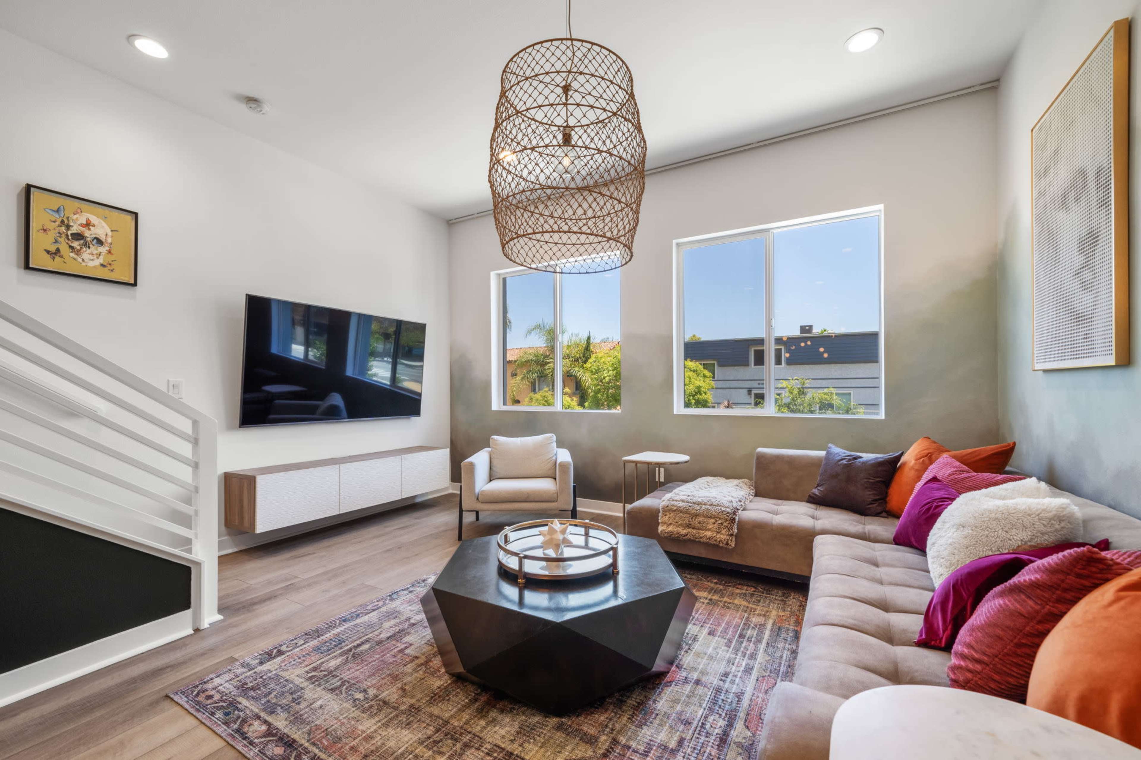 A modern living room features a large sectional sofa, a central coffee table, a TV mounted on the wall, and large windows allowing natural light to illuminate the space.