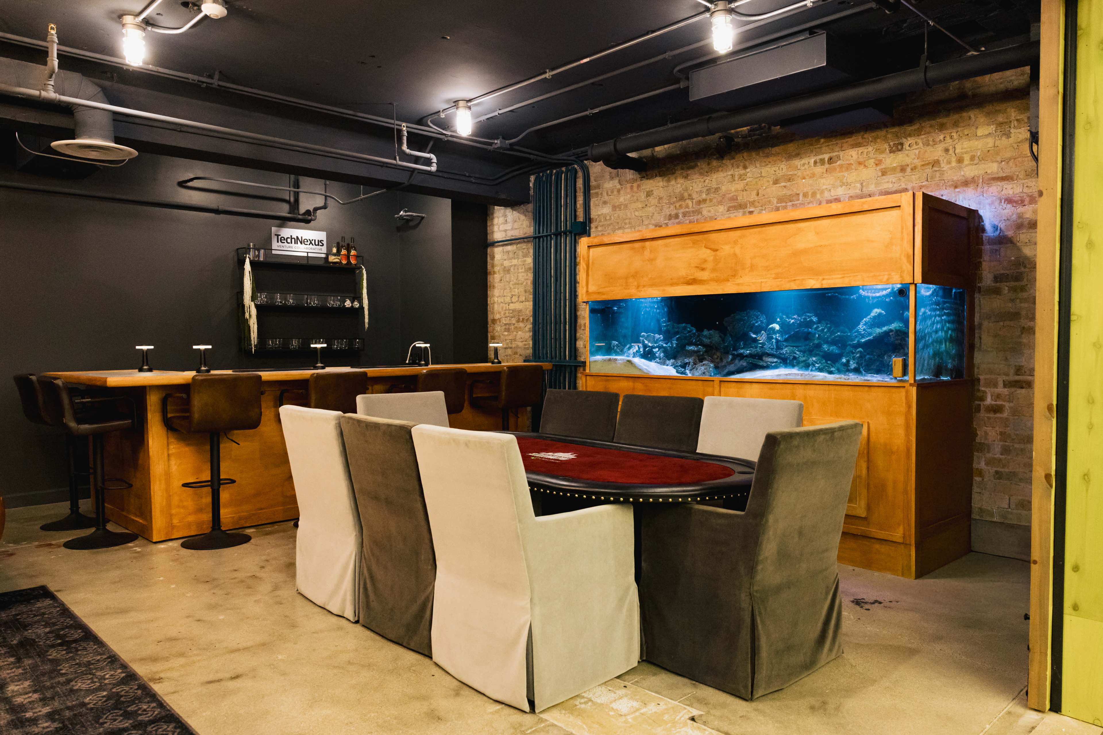 The image shows a dimly lit room featuring a bar area with high stools, a large table surrounded by cushions, and an aquarium built into a wooden cabinet against the wall.